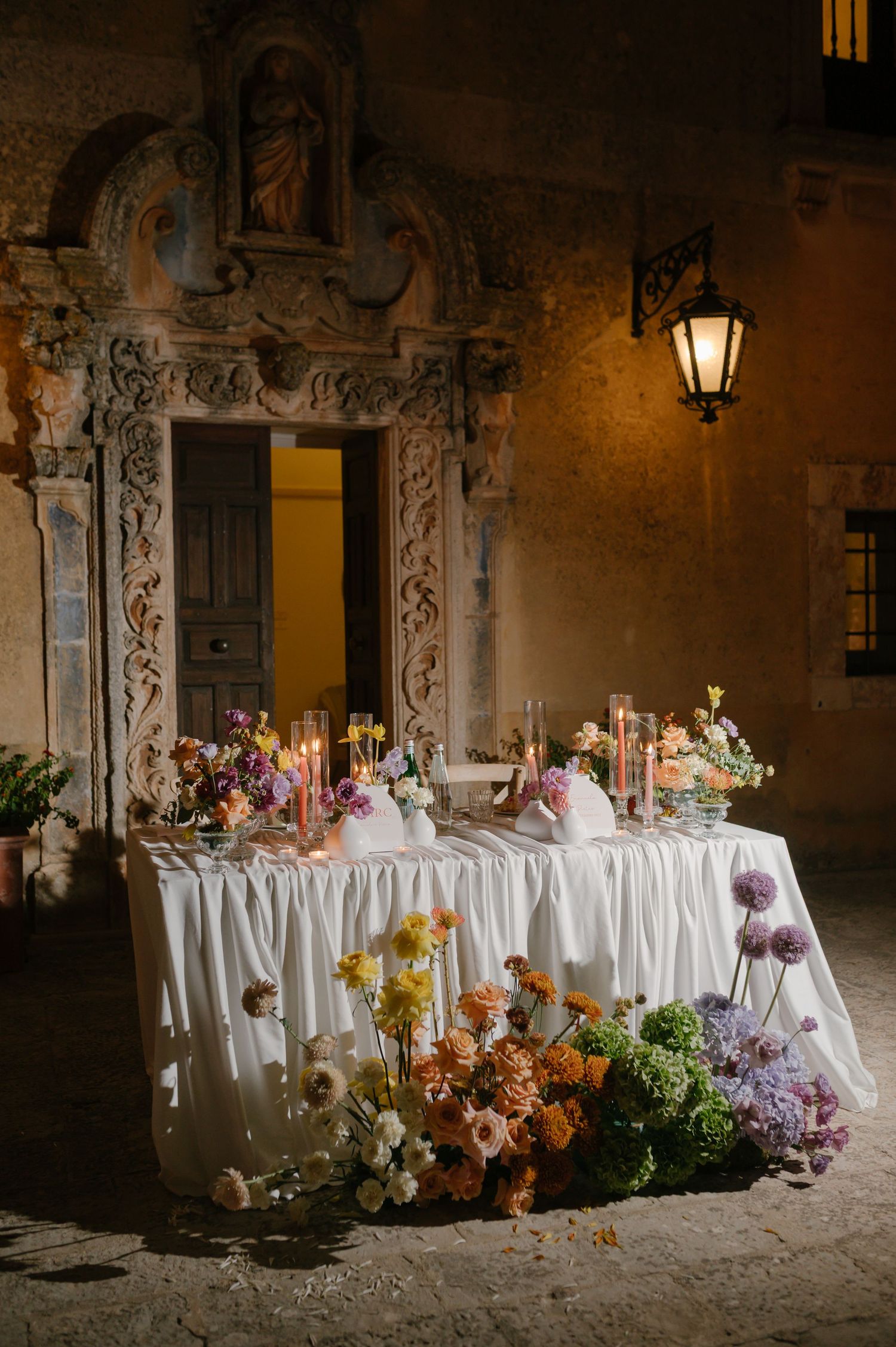 Elegant wedding reception table with floral arrangements and white linens in a stone courtyard.