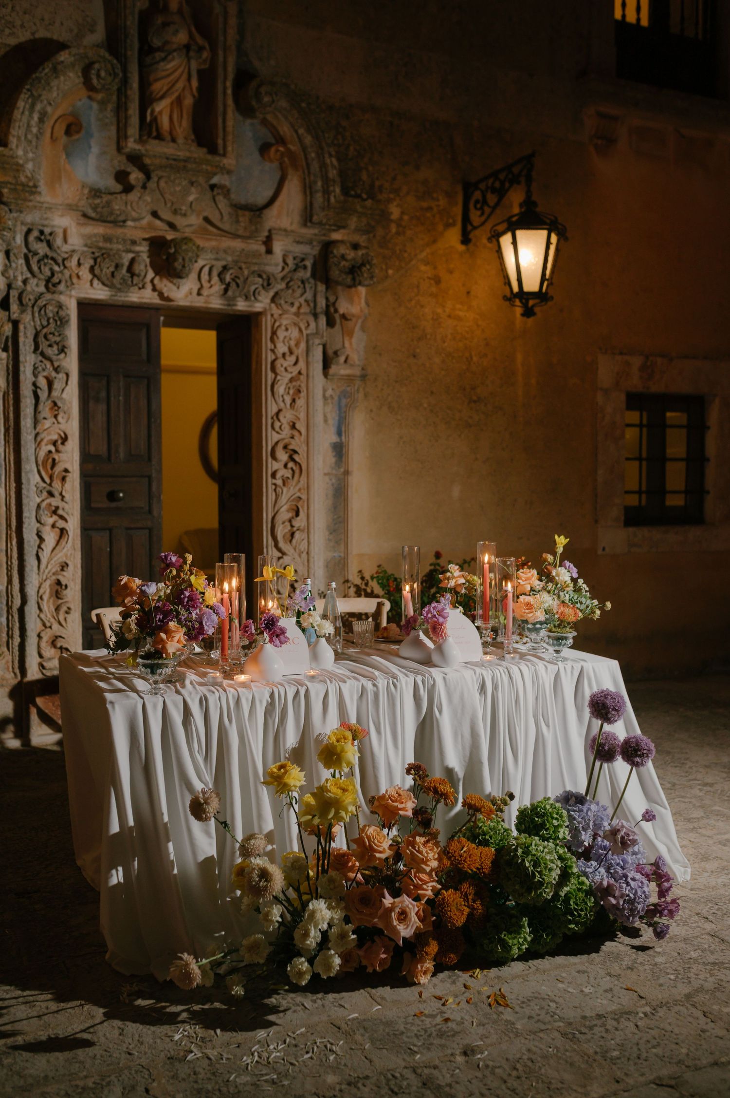 An elegant wedding table with floral arrangements and candles set against an ornate stone doorway lit by a vintage lantern.