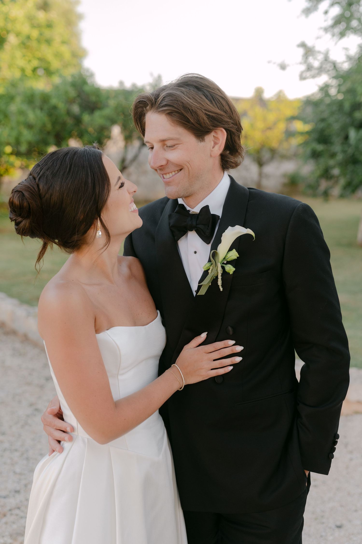 Bride and groom share a romantic moment outdoors, she in a strapless white gown, he in a black tuxedo with floral boutonniere.
