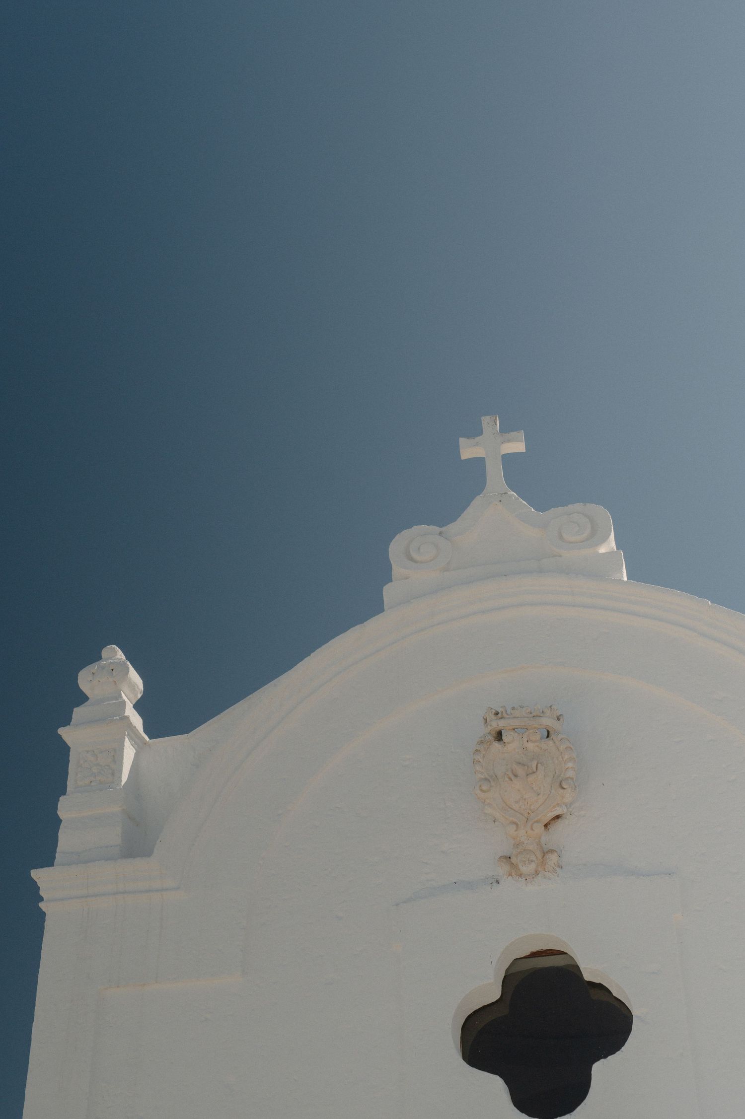 White-washed church facade with ornate cross on top against clear blue sky, Mediterranean architecture.