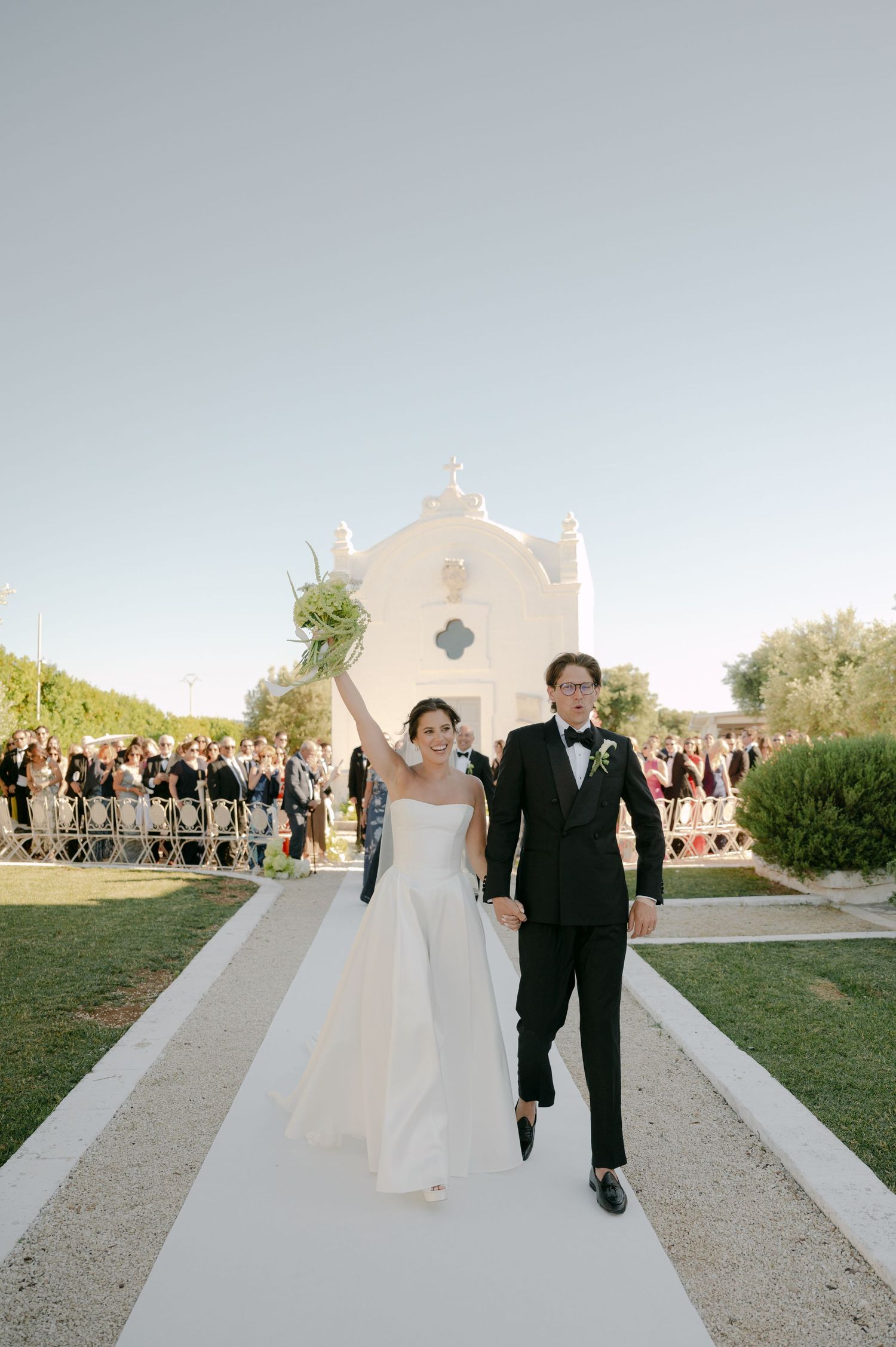 Bride and groom walk down white aisle after ceremony at white chapel wedding venue outdoors.