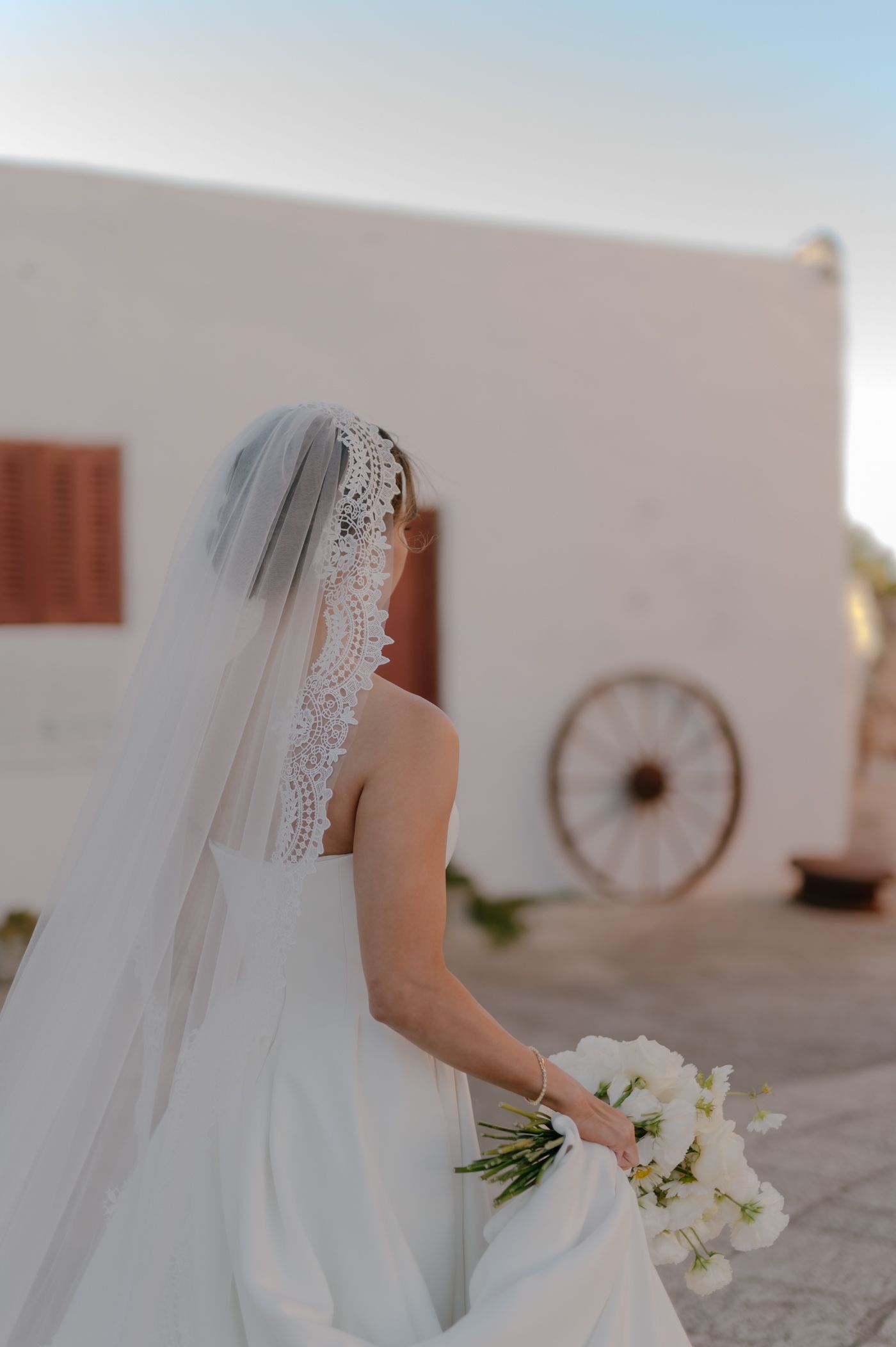 A bride in a white wedding dress and long veil holds a bouquet of white flowers against a rustic wall with a wagon wheel.