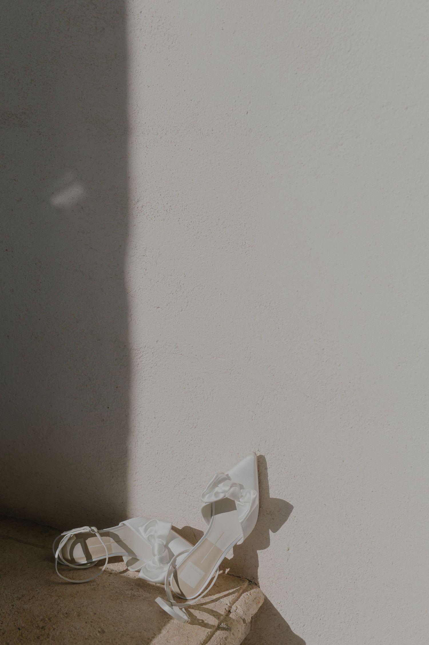 A pair of white sandals lying on a beige floor against a white wall with dramatic shadows.