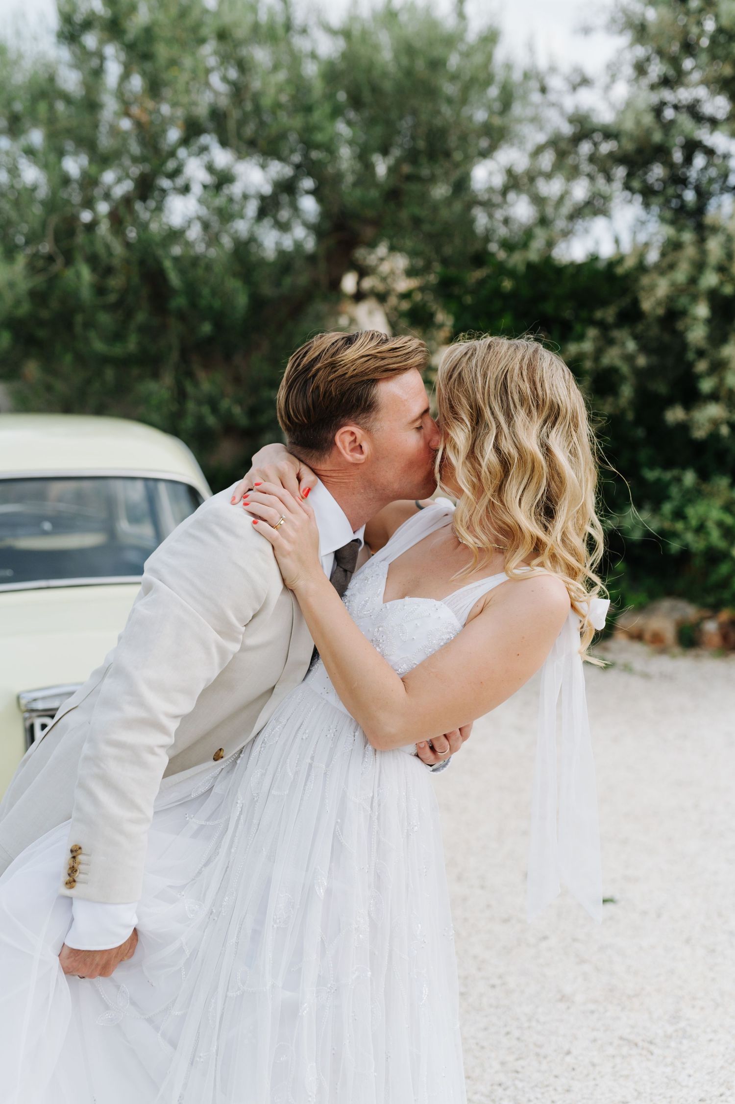 Newlyweds share a passionate kiss beside a classic car on their wedding day.