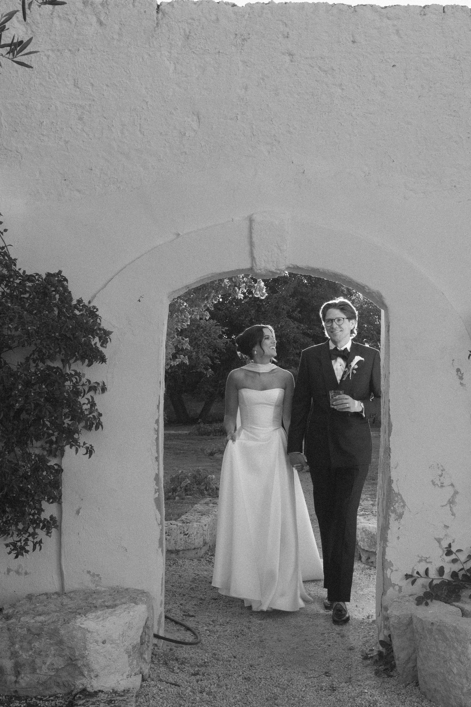 Black and white photo of a bride and groom walking through a stone archway on their wedding day.