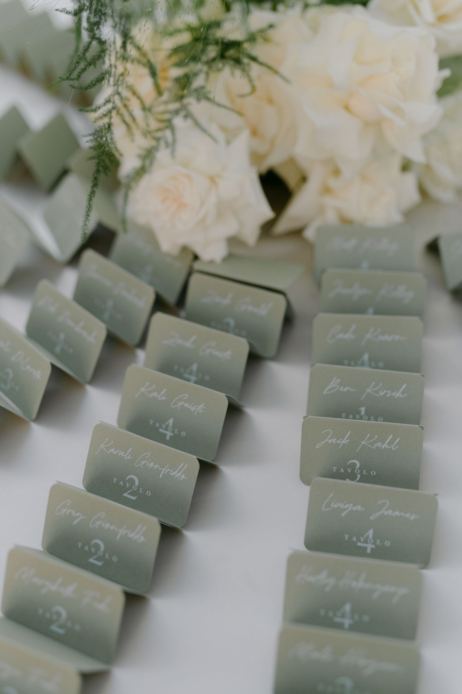 Sage green escort cards with white calligraphy arranged on a table beside white floral arrangements for a wedding reception.