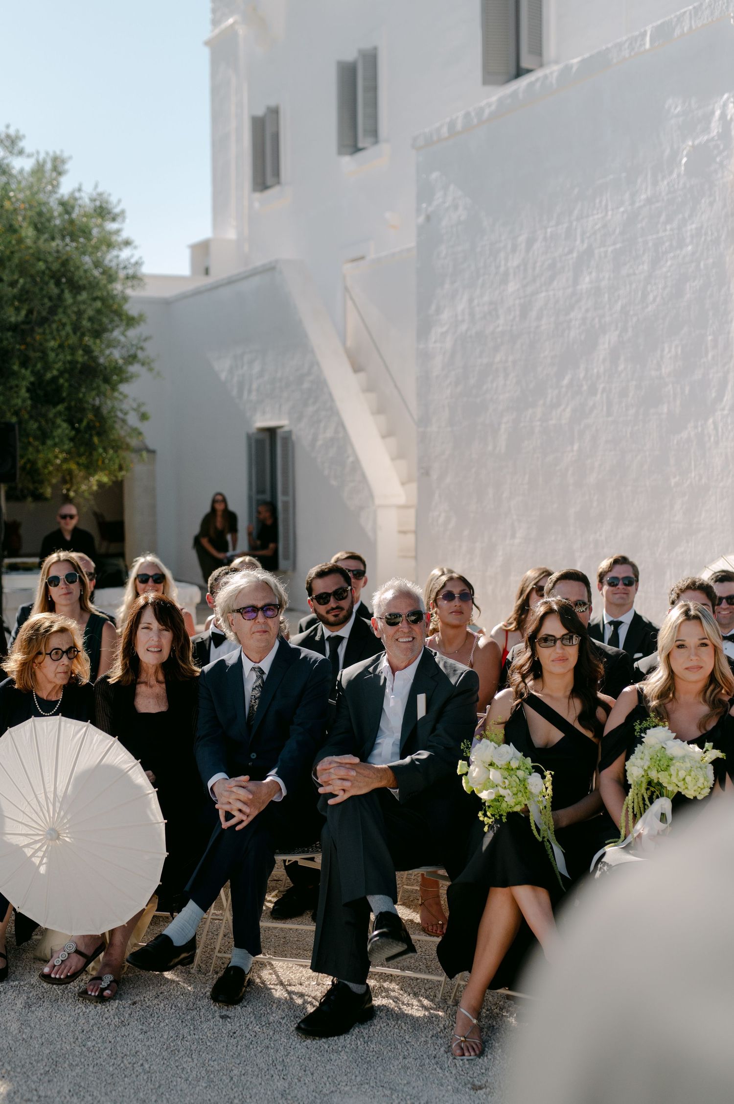 Wedding guests dressed in black seated outdoors at a sunlit ceremony beside a white Mediterranean building.