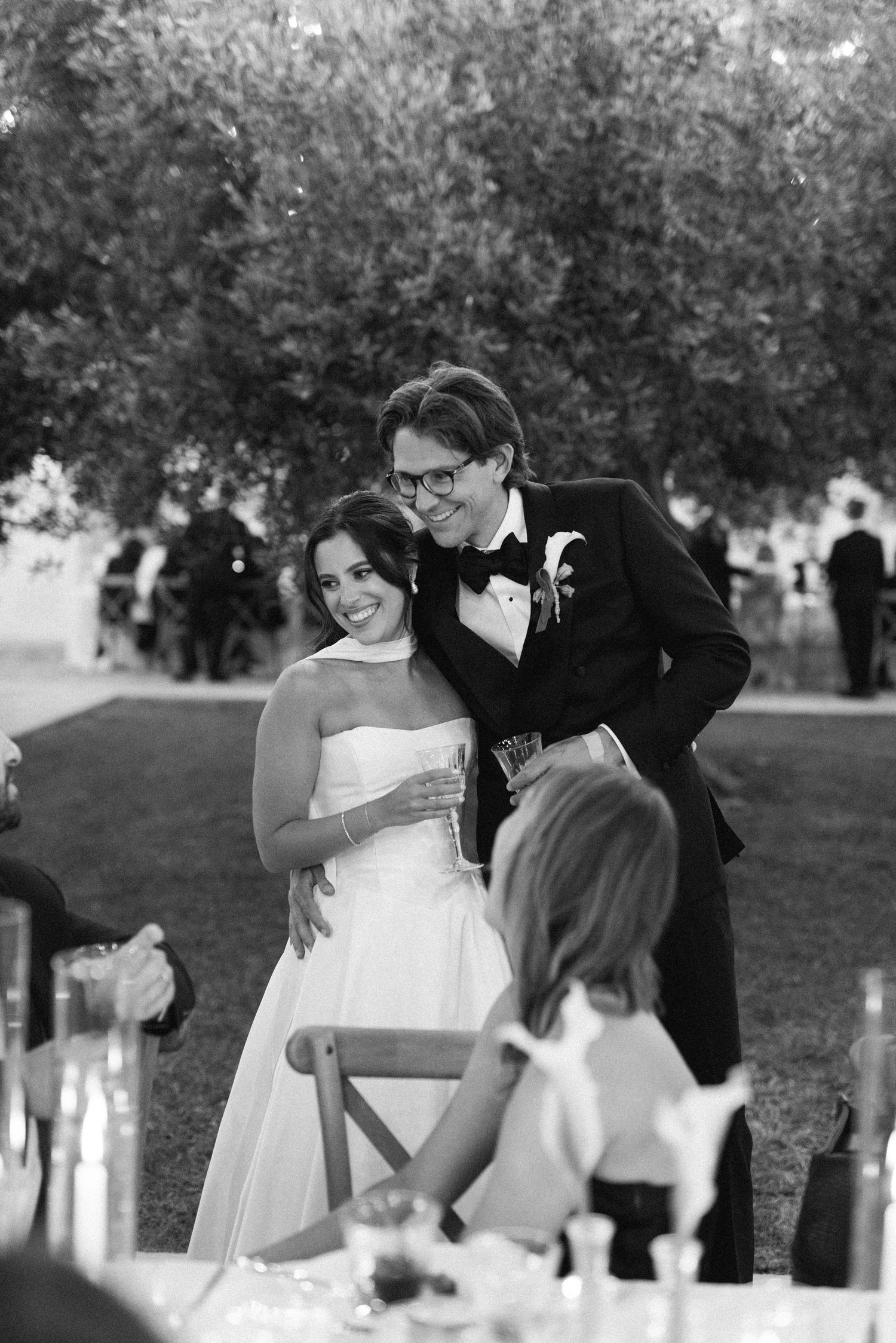 Black and white photo of a smiling bride and groom embracing at their outdoor wedding reception.