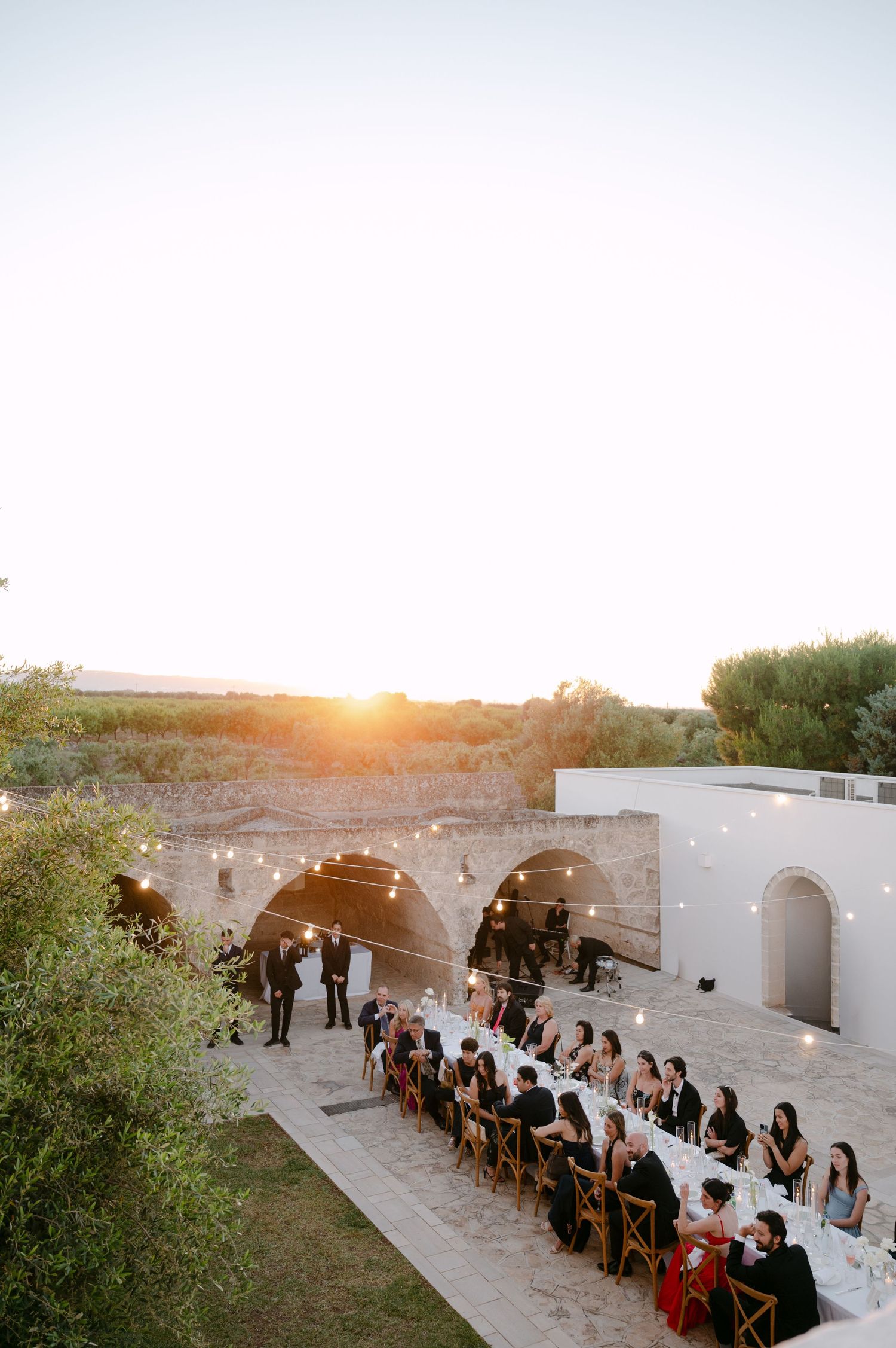 Outdoor wedding reception at sunset with long dining table, string lights, and stone arches at a Mediterranean venue.