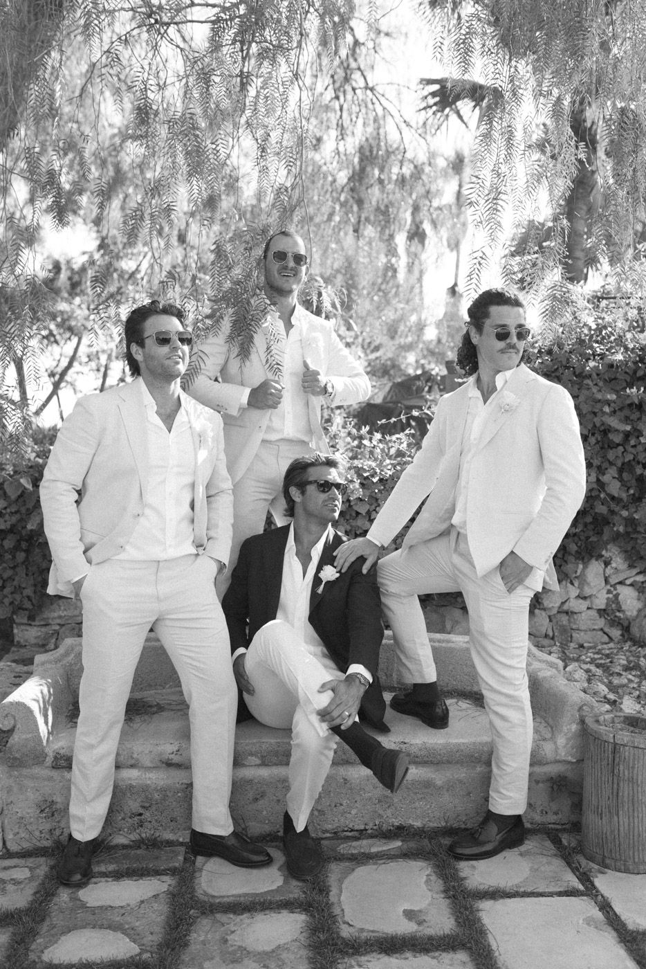 Groomsmen in white suits relaxing on wooden bench under willow trees.