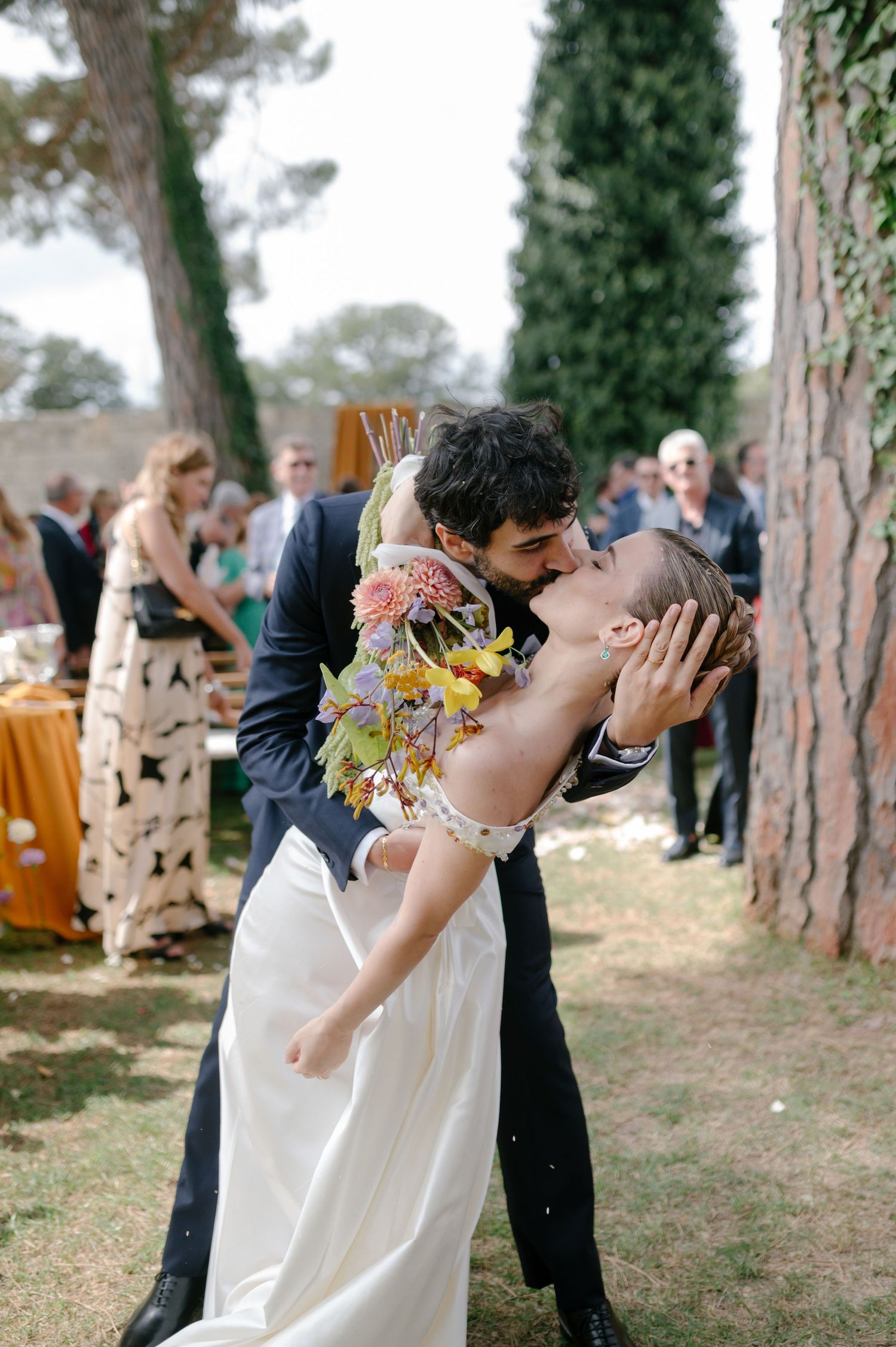 A romantic outdoor wedding moment with a couple sharing a kiss among trees, with the bride wearing a flowing white dress.