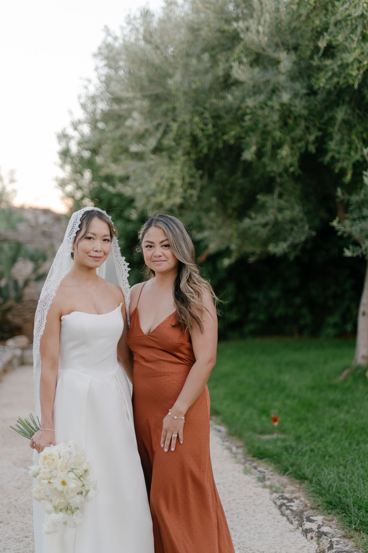 Two people pose together outdoors, one wearing a white gown and one in a rust-colored dress against a natural backdrop.