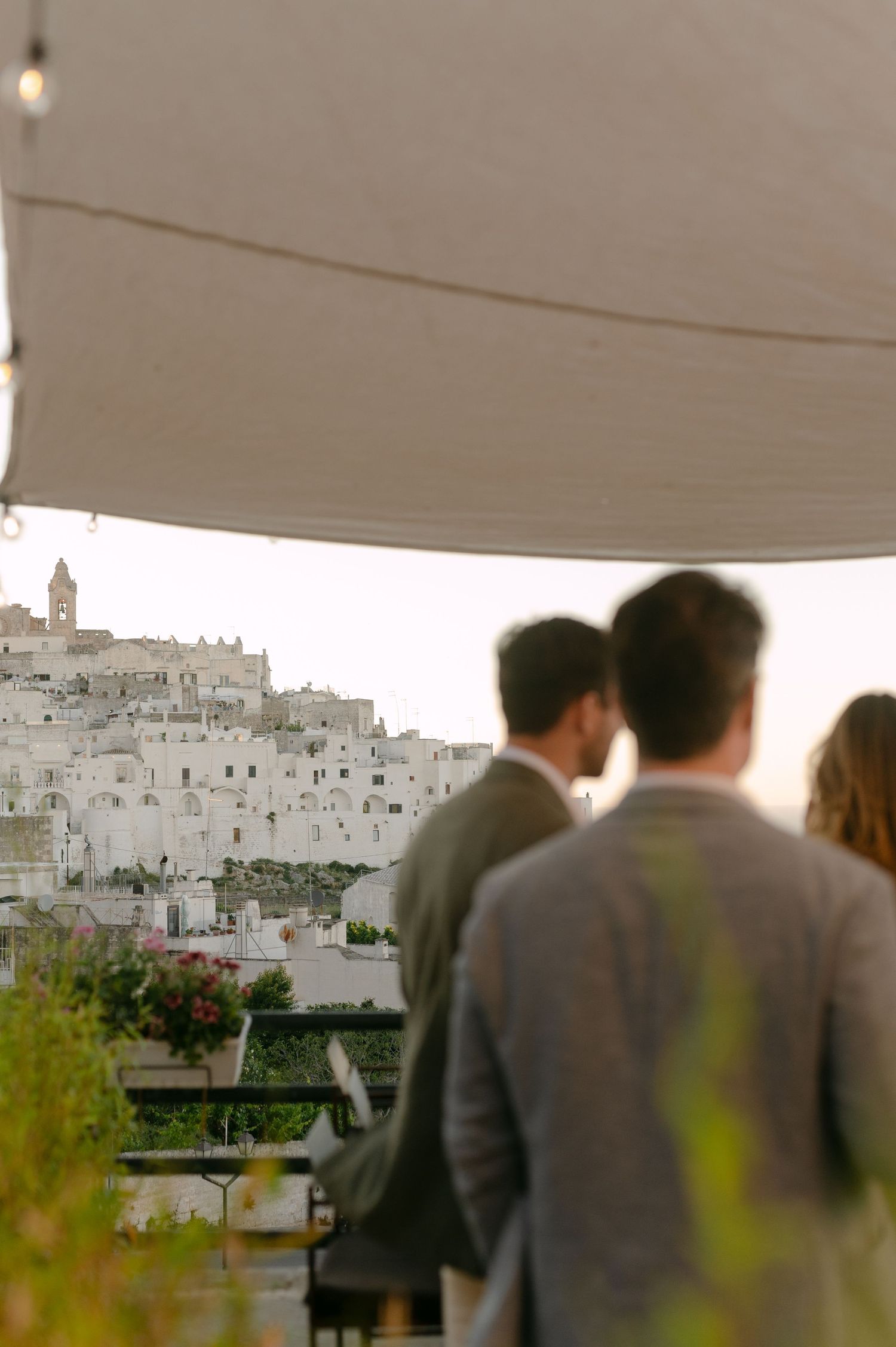 Couple admiring the stunning white hilltop town of Ostuni, Italy, from a terrace under a white canopy.