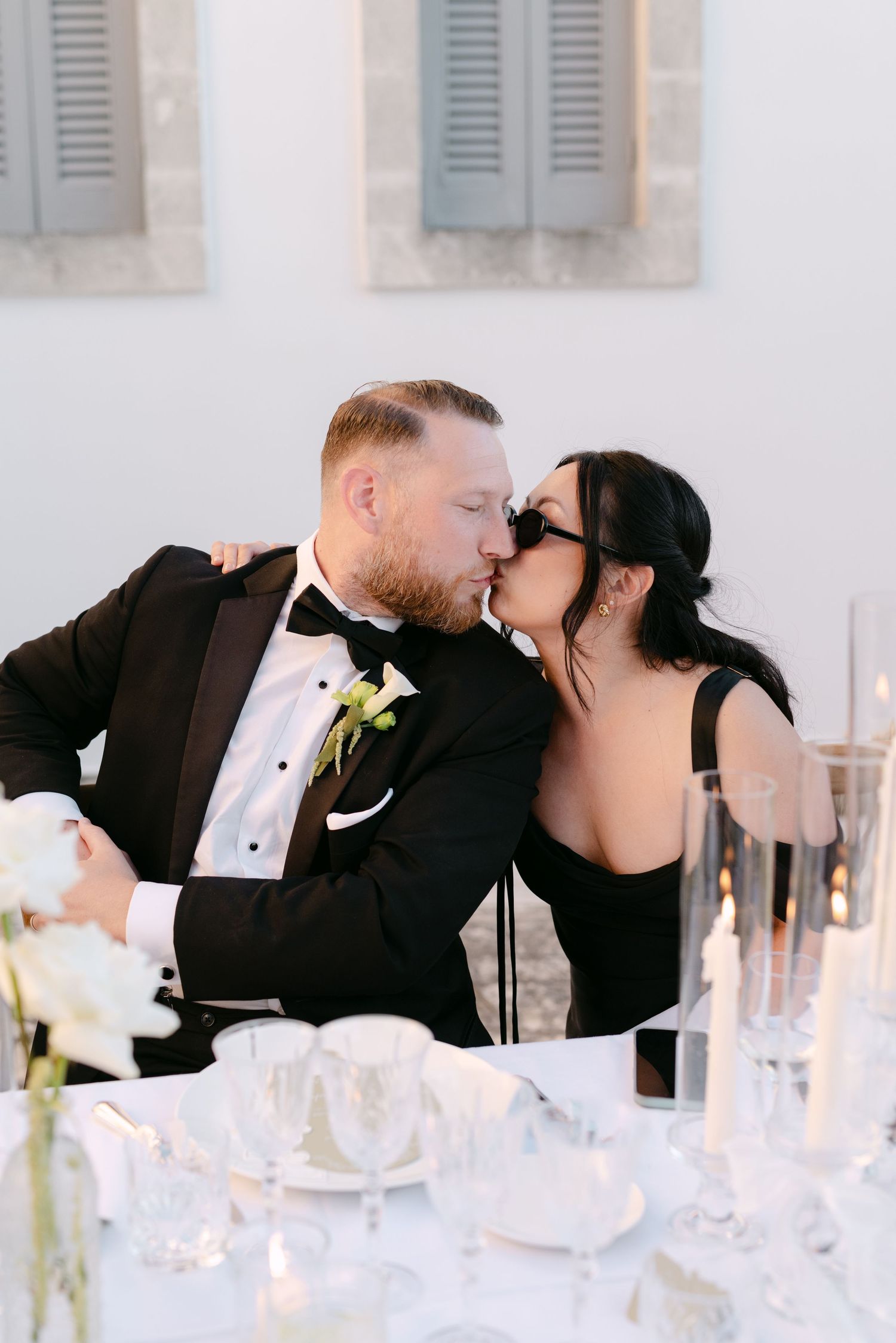 A groom in a black tuxedo and bride in a black dress share a kiss at their elegant wedding reception table.