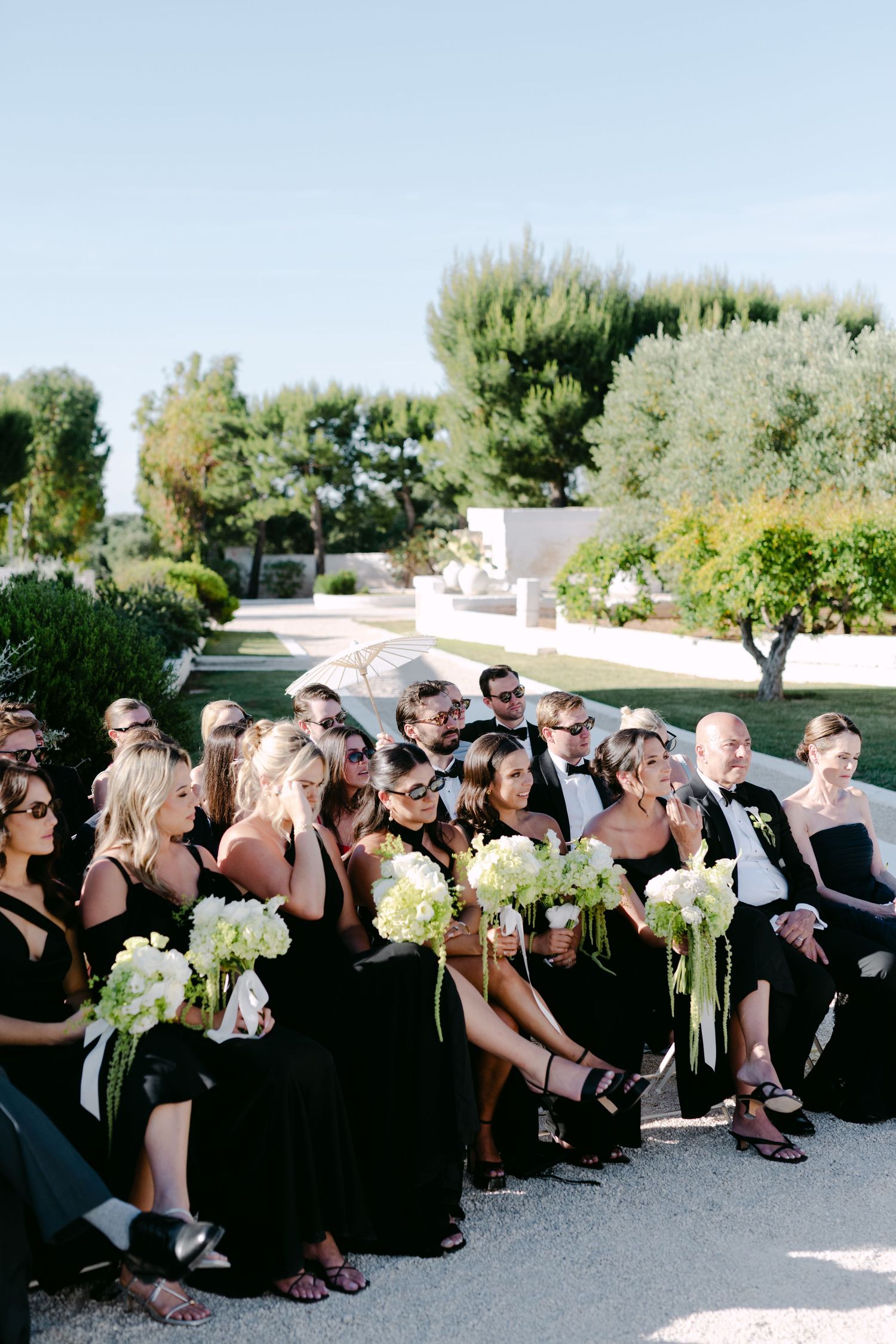 Wedding party in black attire seated outdoors, bridesmaids holding white floral bouquets at an elegant garden ceremony.