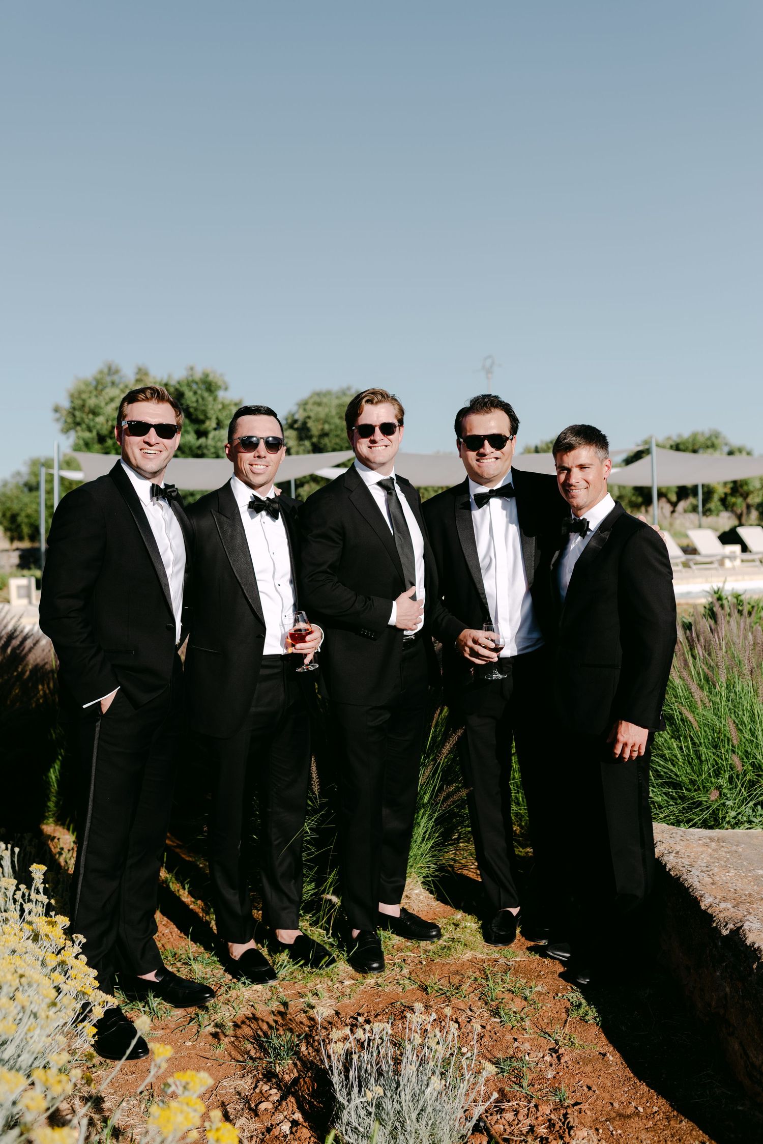 Five groomsmen in black tuxedos and sunglasses posing outdoors on a sunny wedding day.