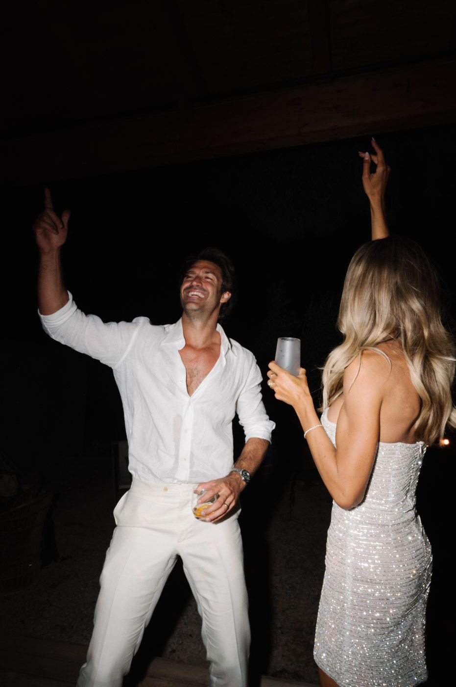 Couple dancing energetically in white attire at a dimly lit reception.