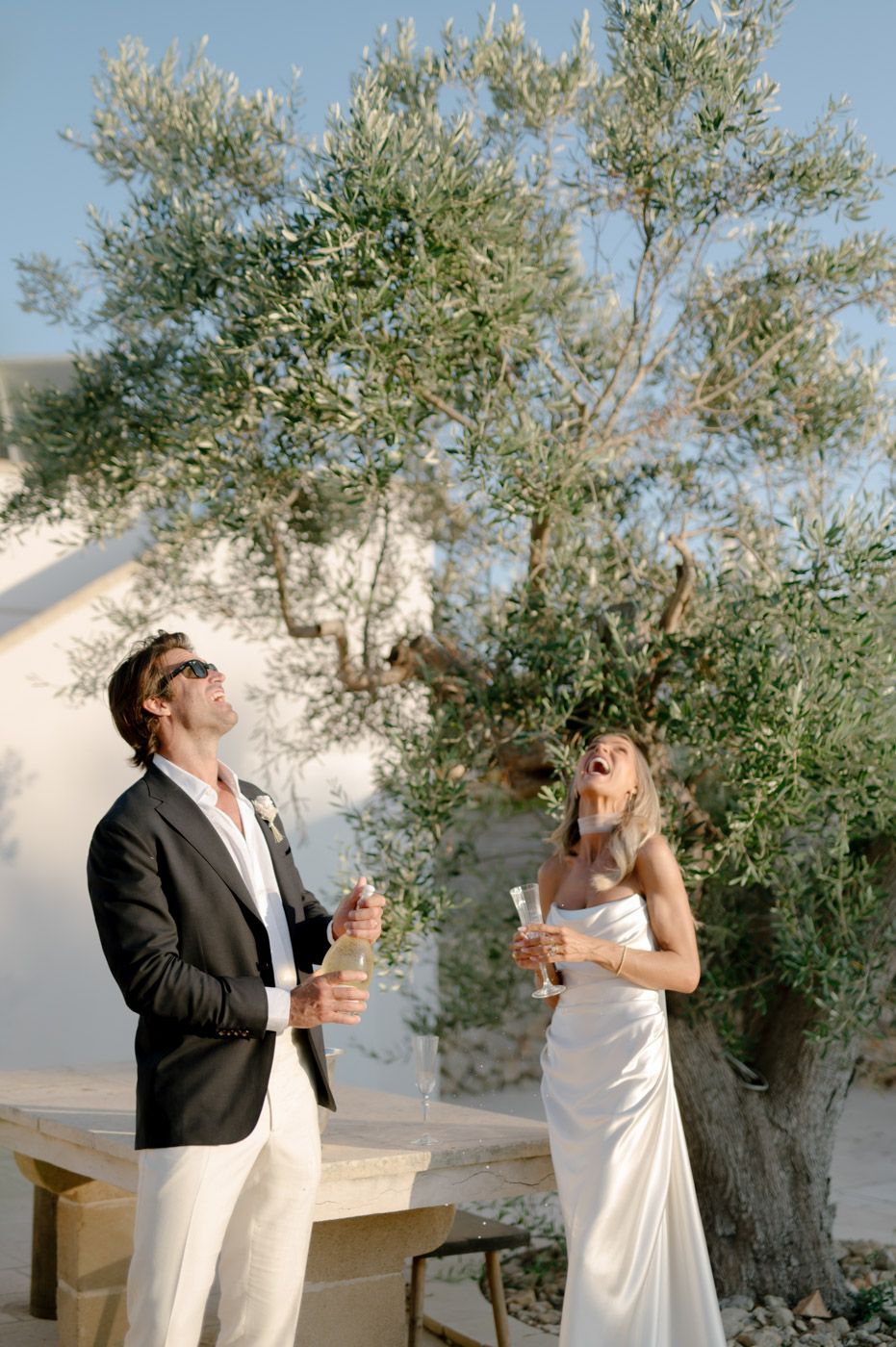 Couple enjoying drinks under olive trees at an outdoor celebration.
