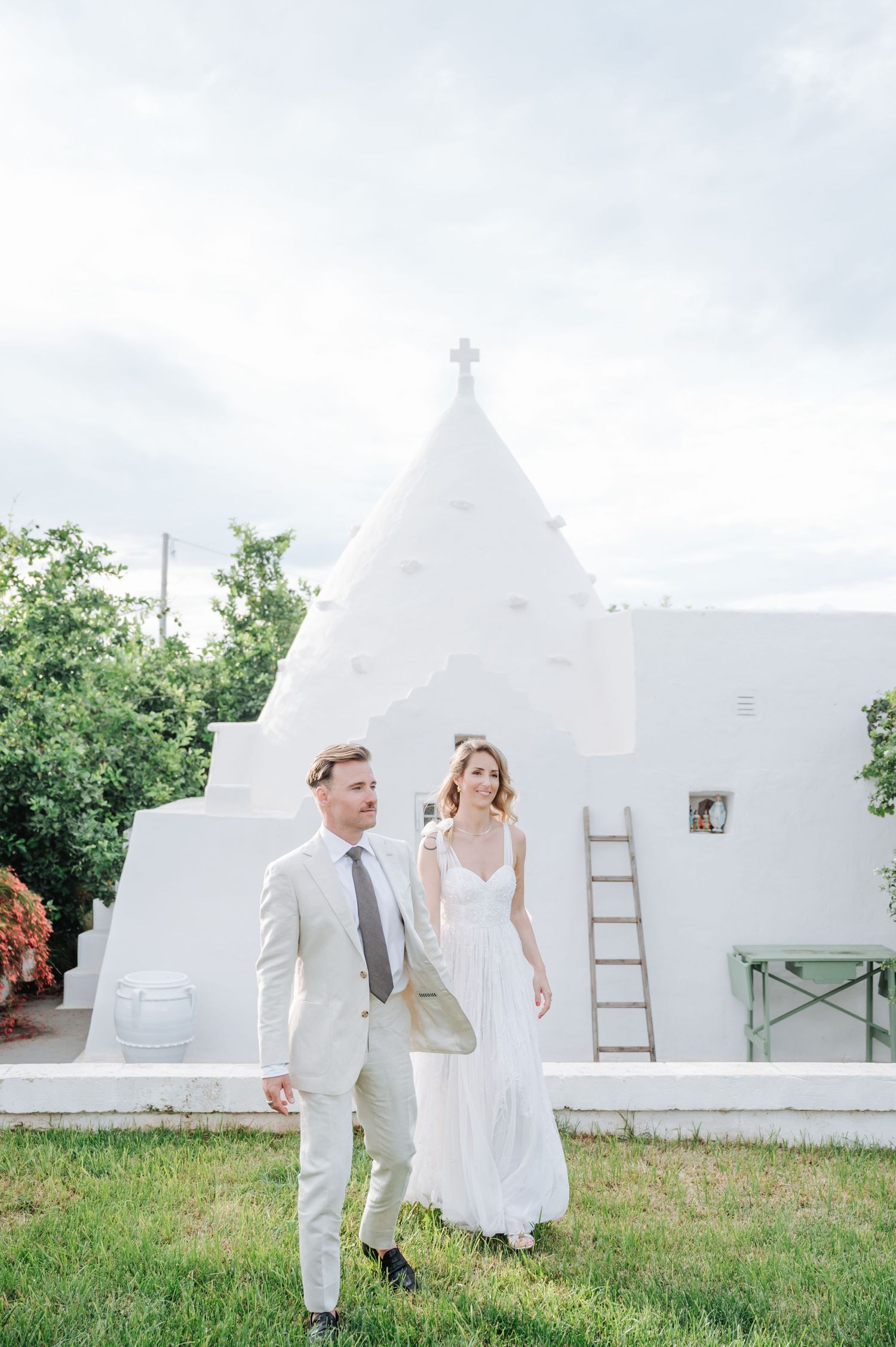 A wedding couple poses in front of a white chapel with a cross on top.