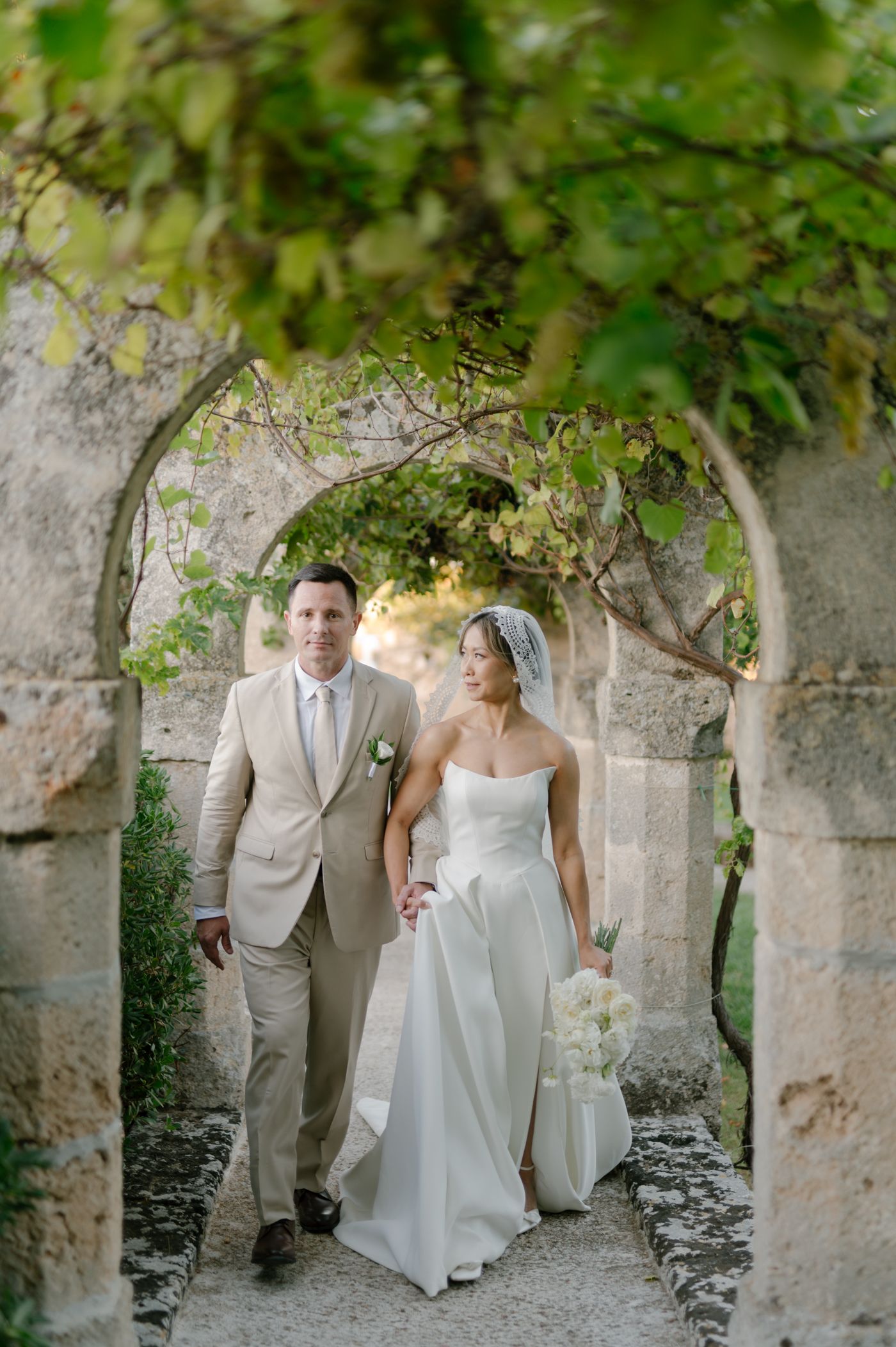 A couple in wedding attire walks through a romantic stone archway covered in green vines and foliage.