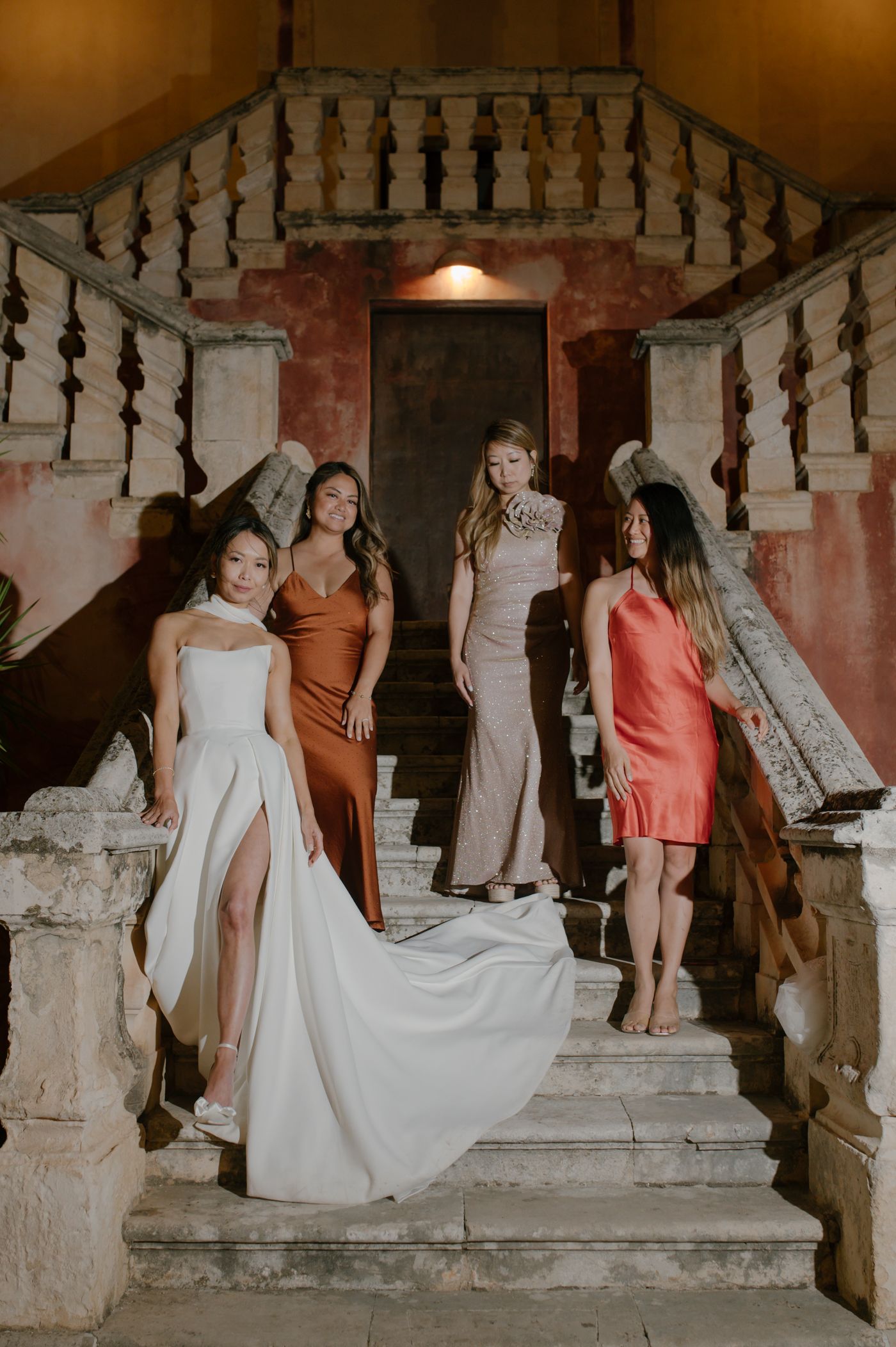 A group stands elegantly on vintage stone stairs in formal attire including flowing gowns in white, rust, and coral colors.