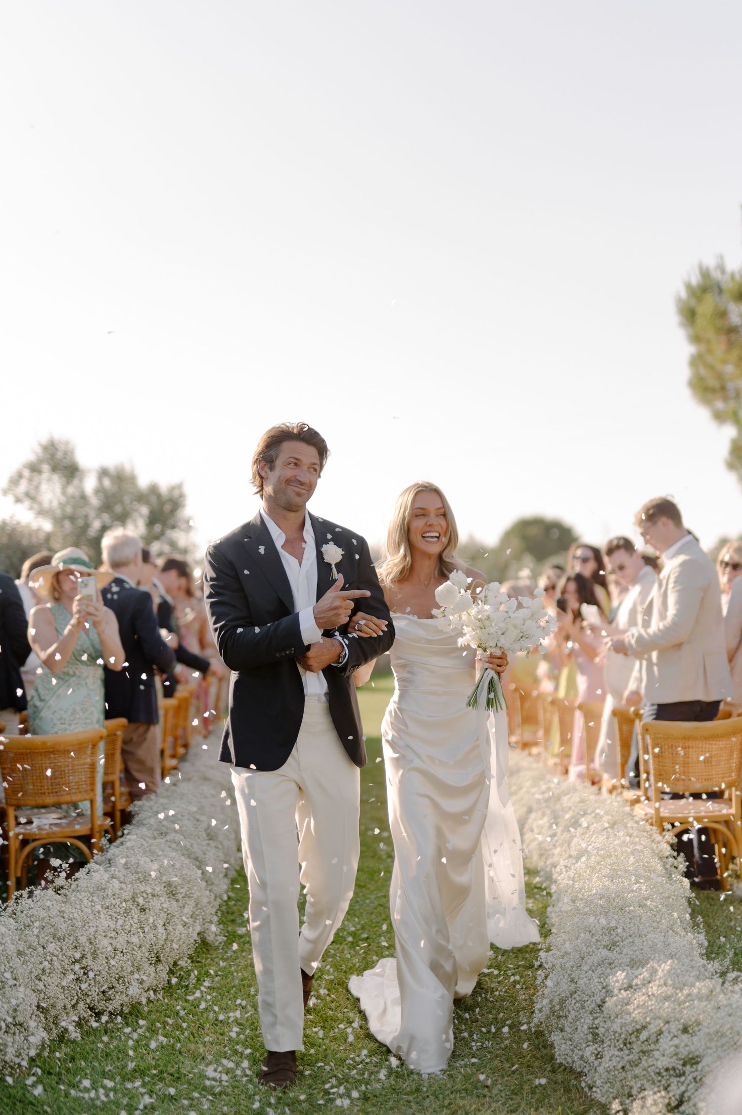 Wedding guests throw confetti and celebrate outdoors with wooden chairs and natural scenery in the background.