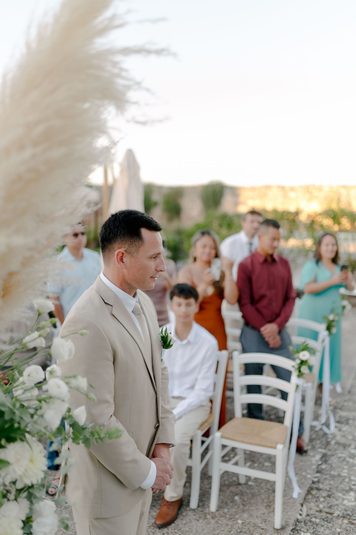 Outdoor beach wedding ceremony setup with white chairs and guests gathered at sunset.
