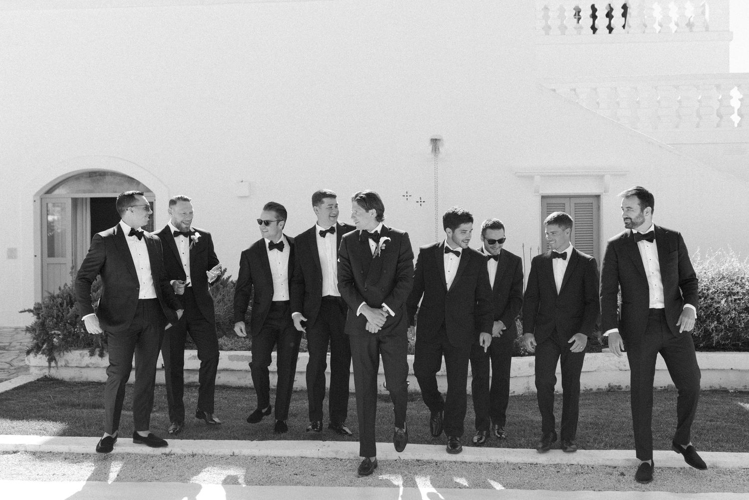 Black and white photo of groomsmen in tuxedos and sunglasses walking confidently together outside a white building.