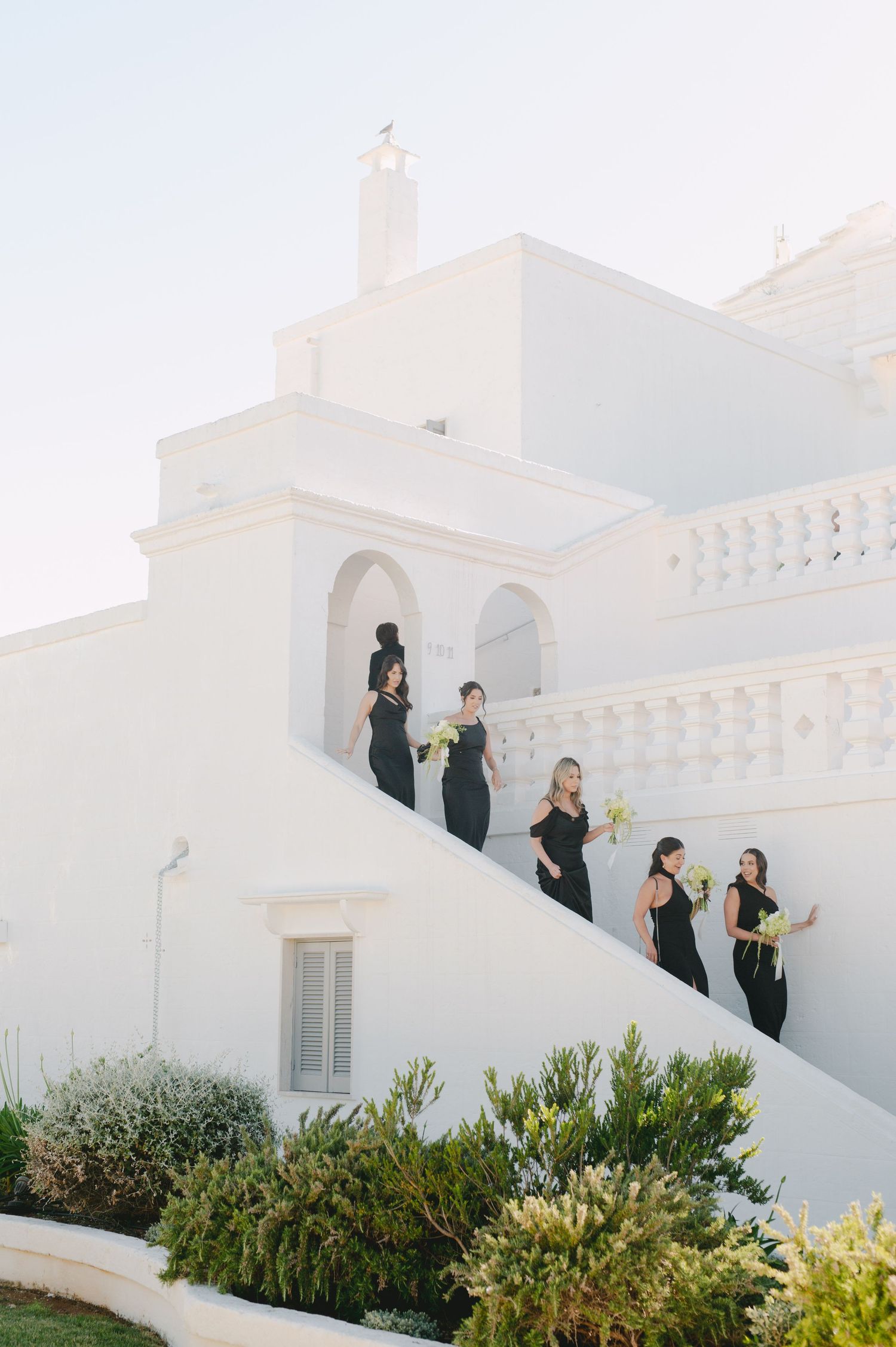Bridesmaids in black gowns holding bouquets descend white Mediterranean villa stairs in golden sunlight.