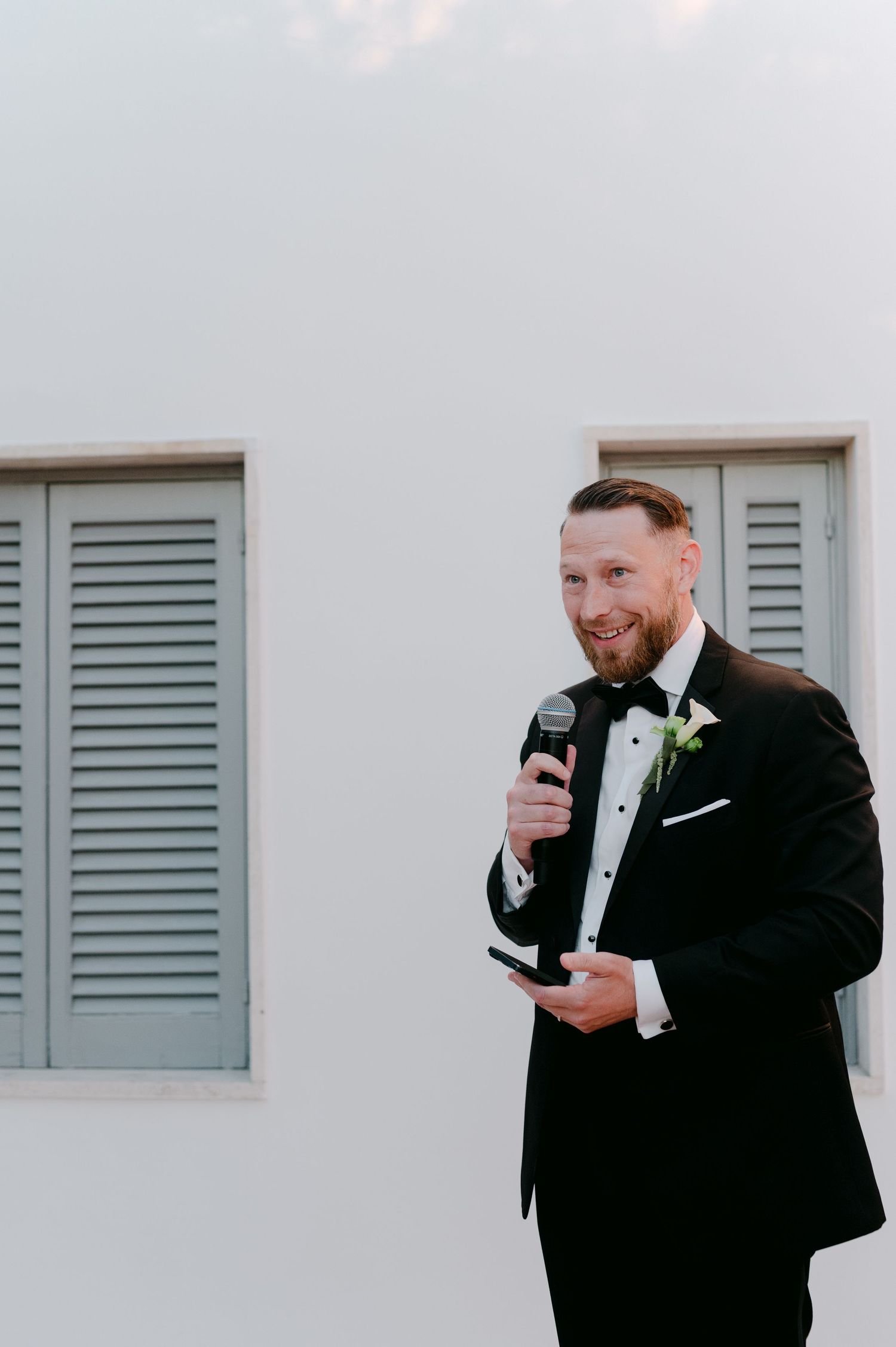 Groom in black tuxedo with boutonniere speaks into microphone at wedding reception outdoors.