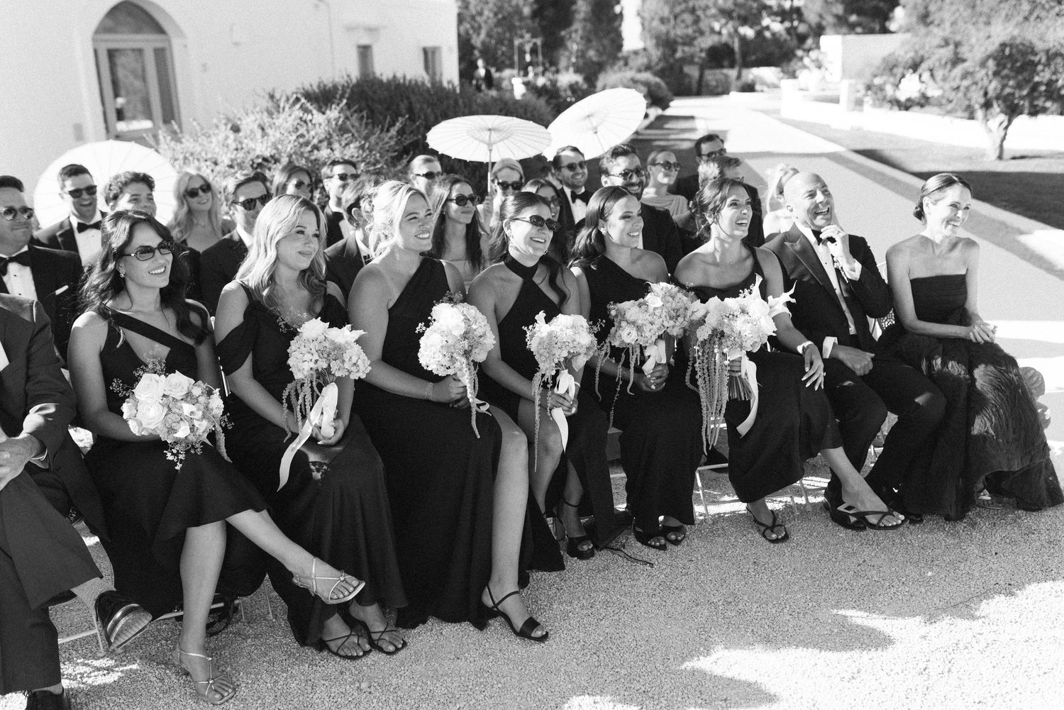 Black and white photo of wedding party in black attire holding white bouquets outside a church.