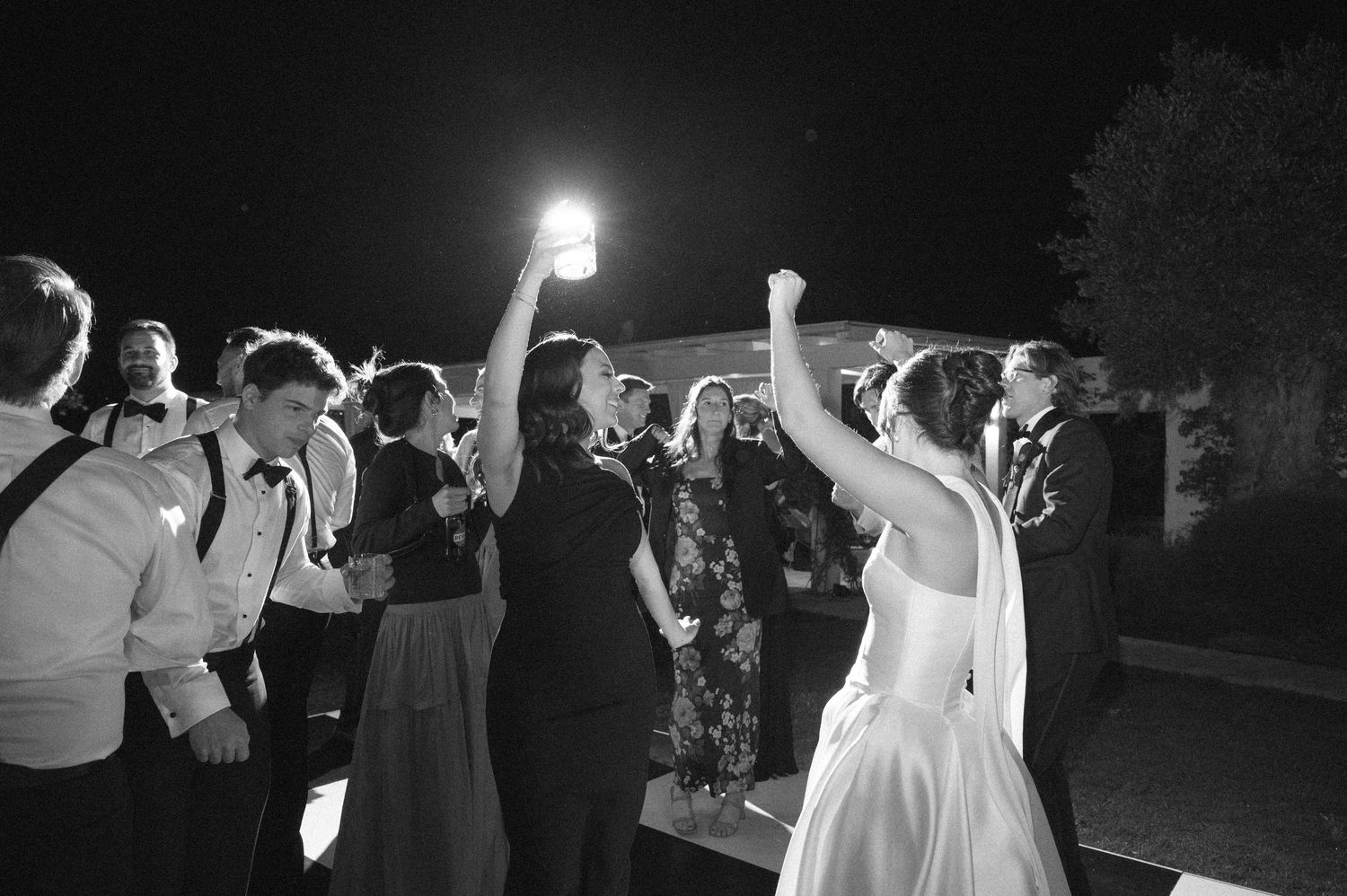 Black and white photo of a bride and guests dancing outdoors at night during a wedding reception celebration.