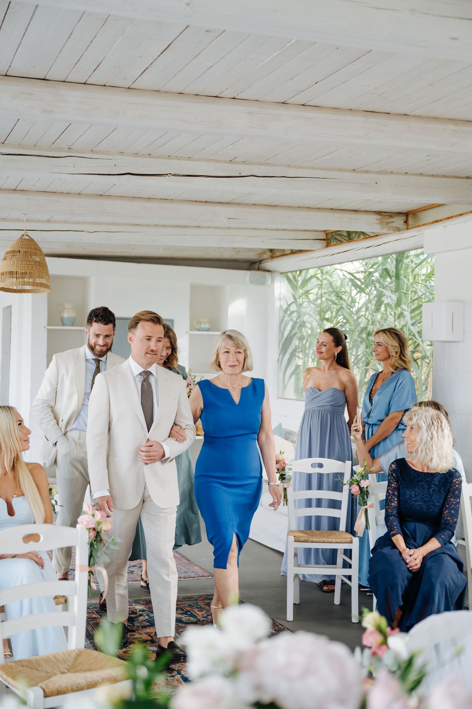 Wedding guests gather in an airy white room with wooden ceiling beams and natural light.