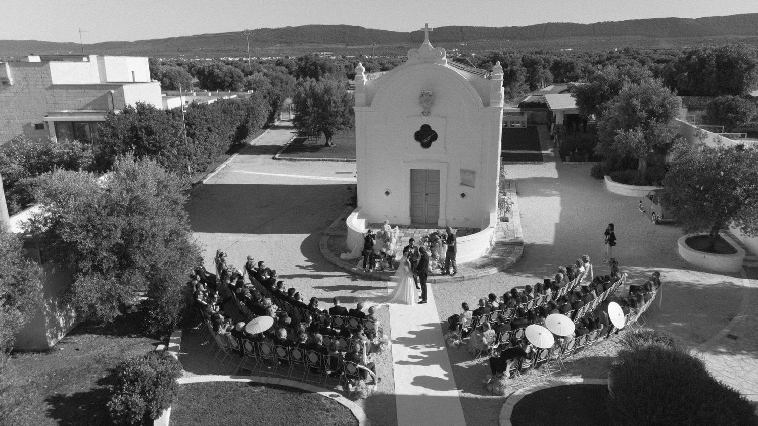 Aerial black and white photo of an outdoor wedding ceremony at a small white chapel surrounded by trees.