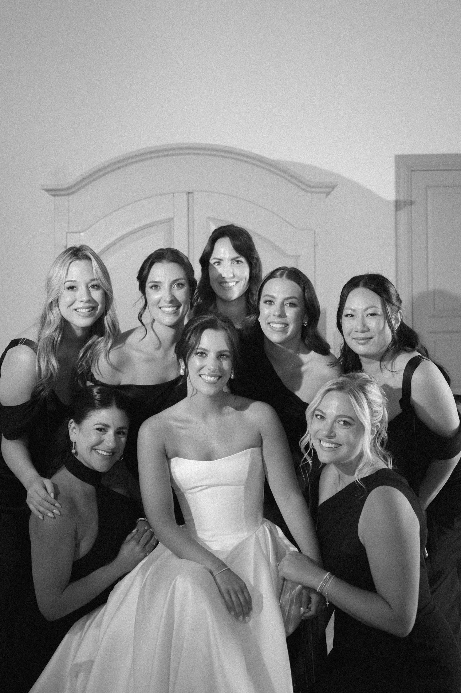 Black and white photo of a bride in a strapless gown surrounded by seven bridesmaids in dark dresses.