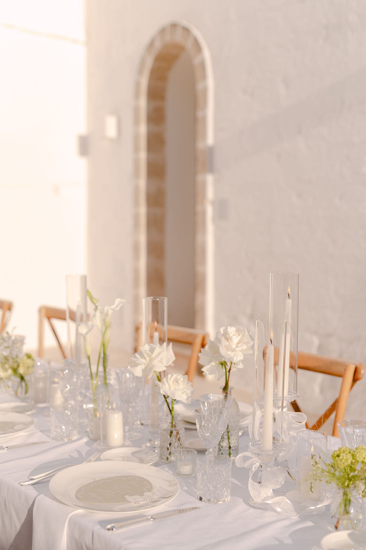 Elegant white wedding table setting with tall candles, white flowers, and glassware on a linen tablecloth.