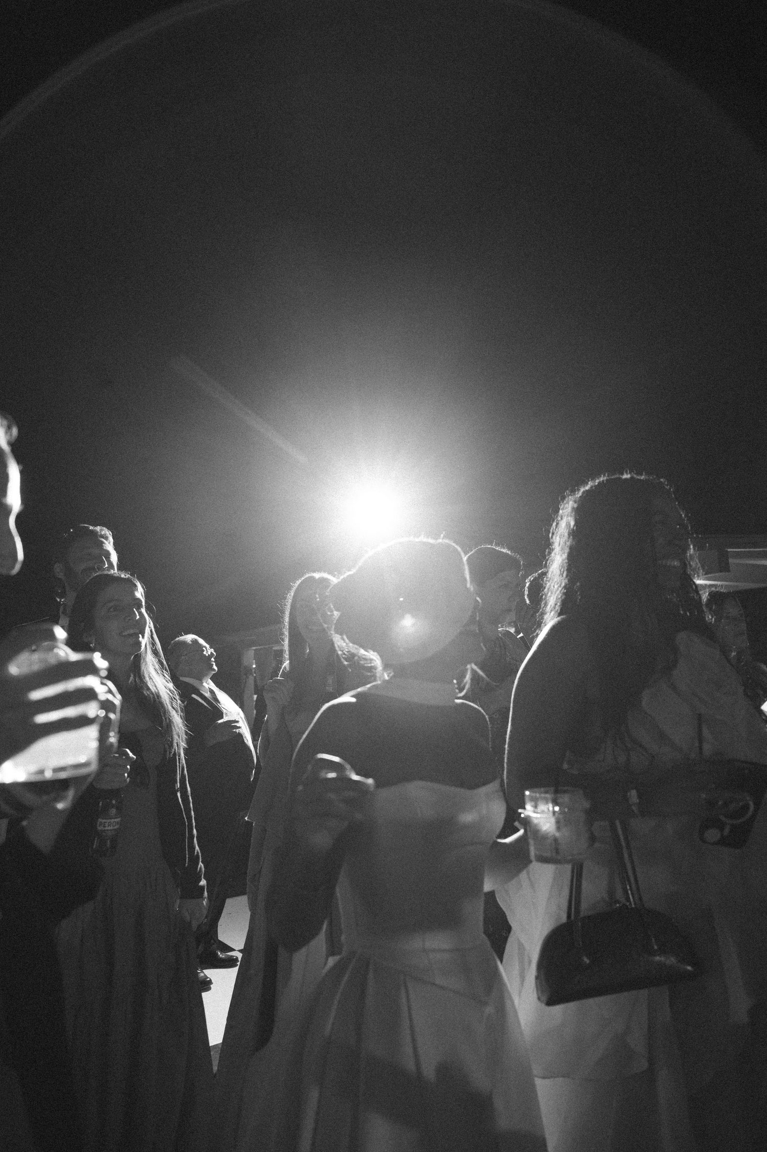 Black and white photo of elegantly dressed guests at a formal evening event, illuminated by a bright spotlight.