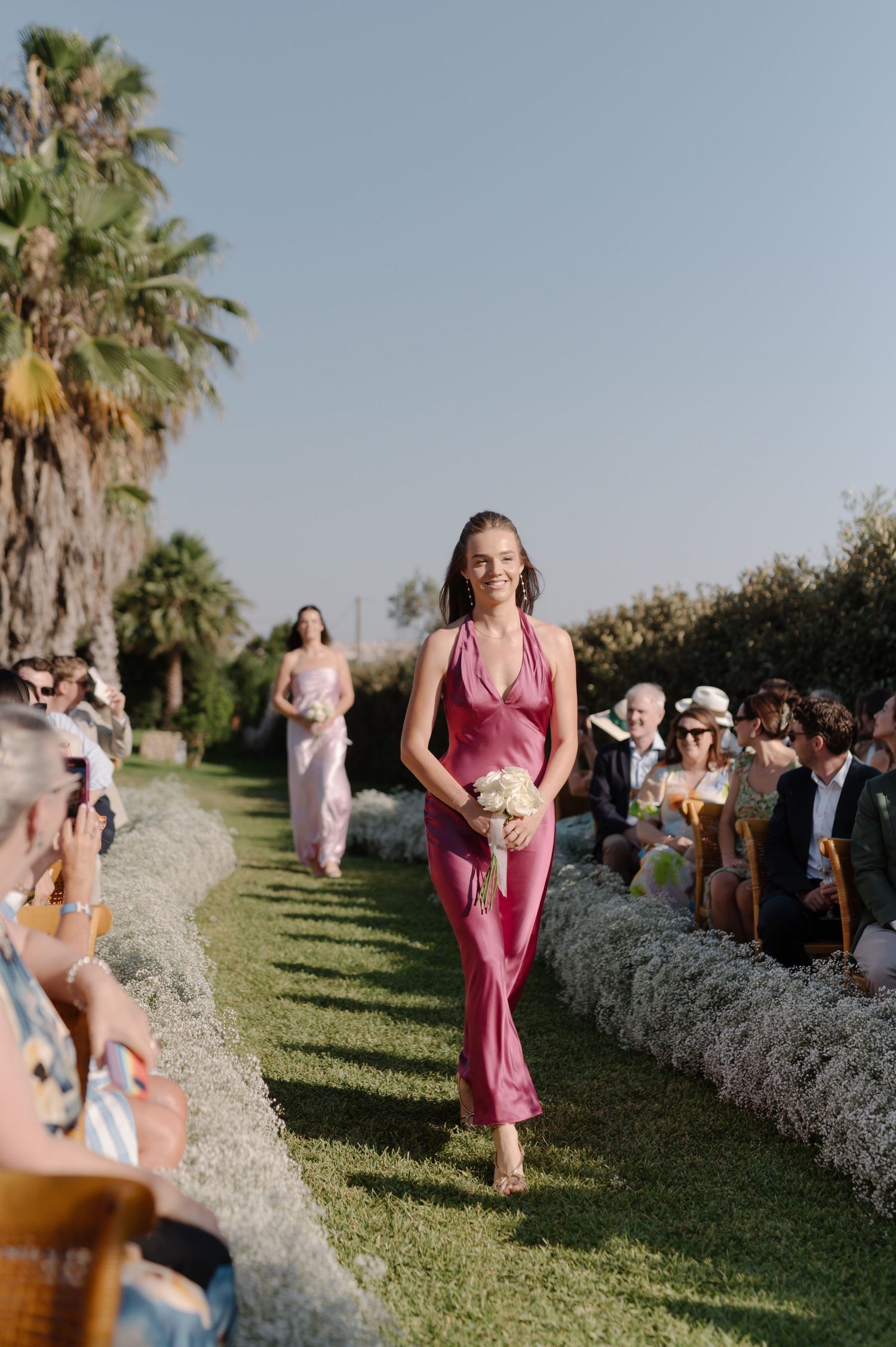 A pink-dressed bridesmaid walks down a grassy outdoor aisle lined with palm trees and seated wedding guests.