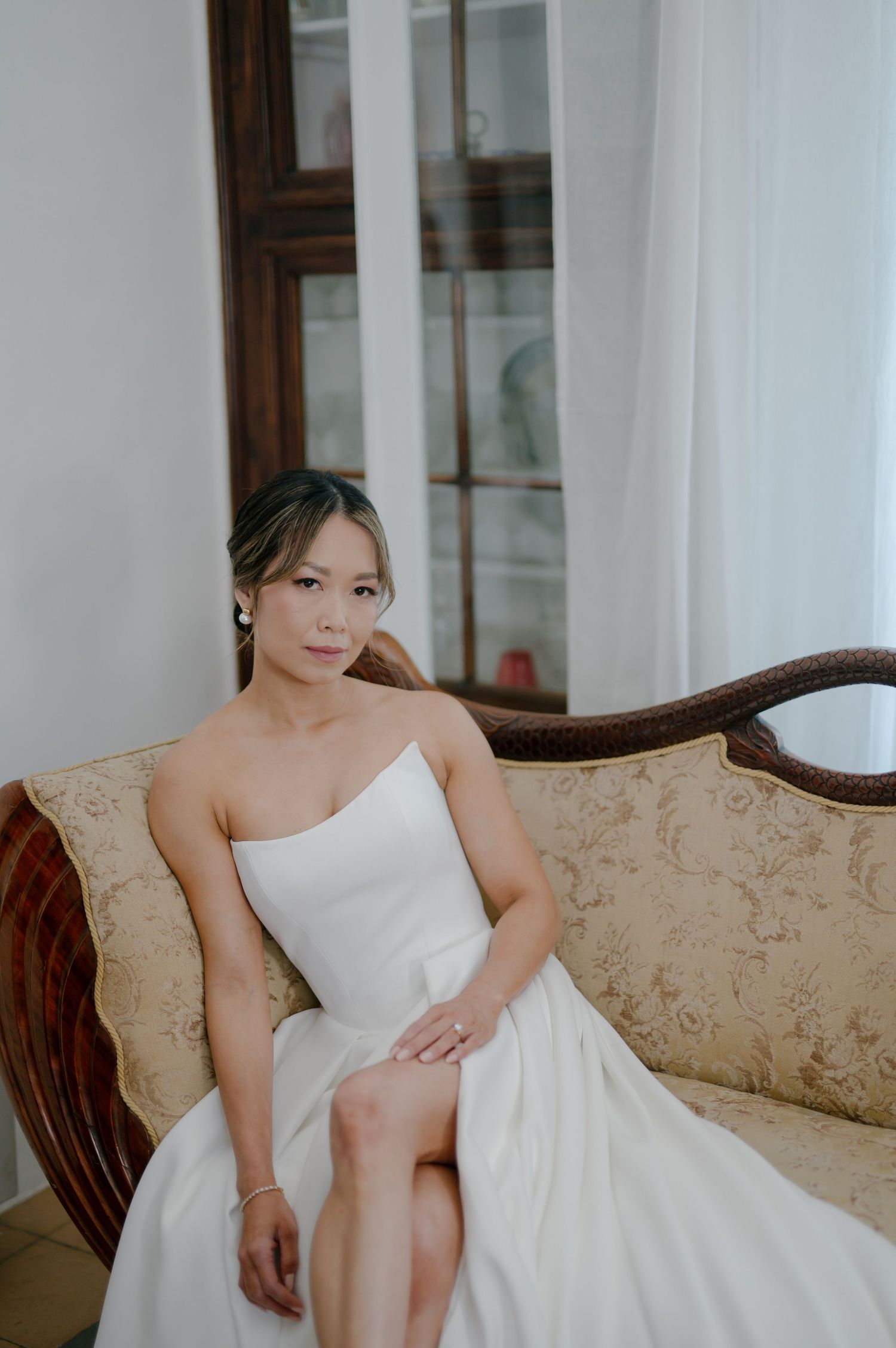 A bride in a white strapless wedding dress sits gracefully on an elegant beige damask sofa in an indoor setting.