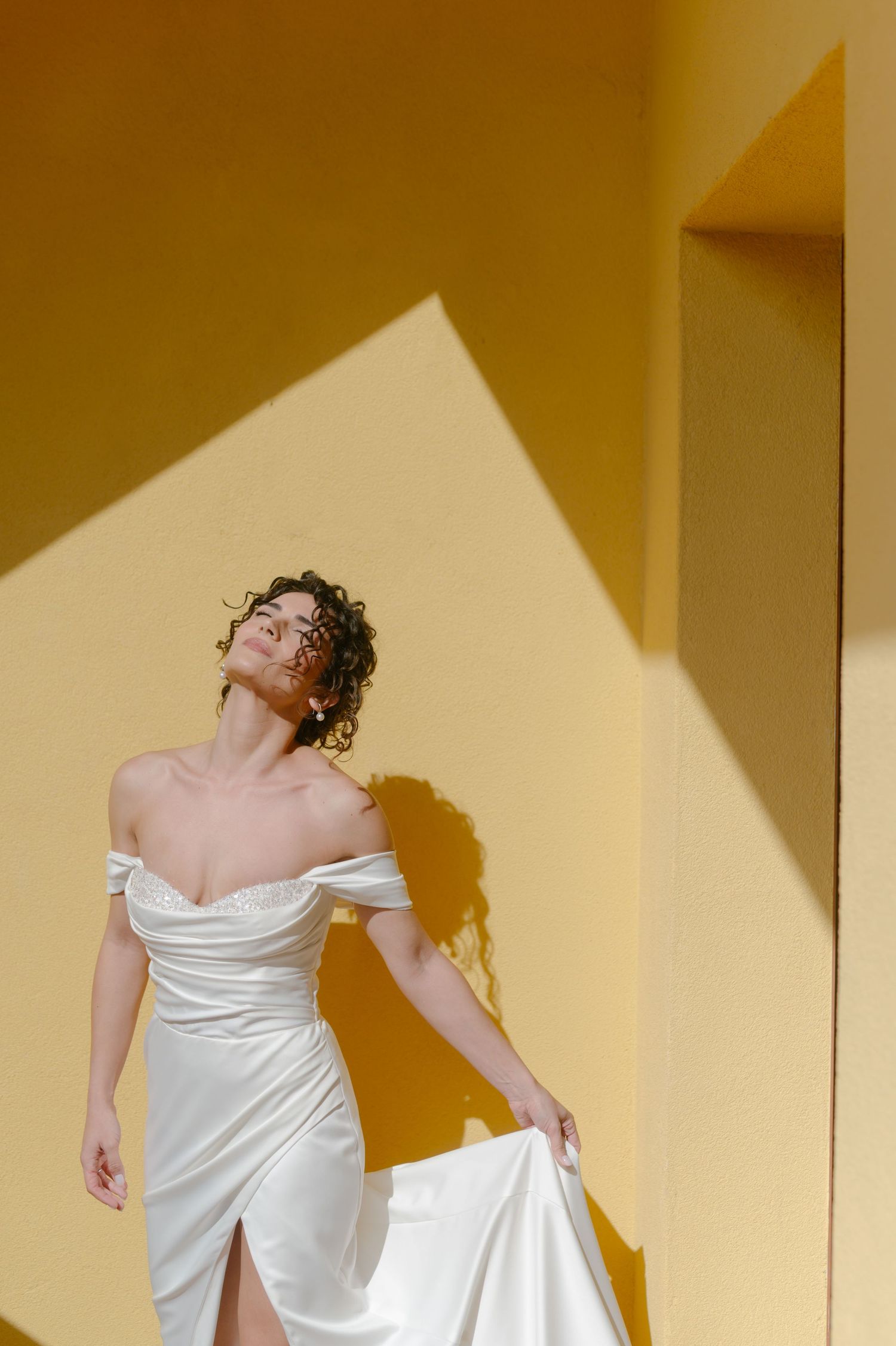 Bride in white off-shoulder wedding dress posing against yellow wall with dramatic shadows.