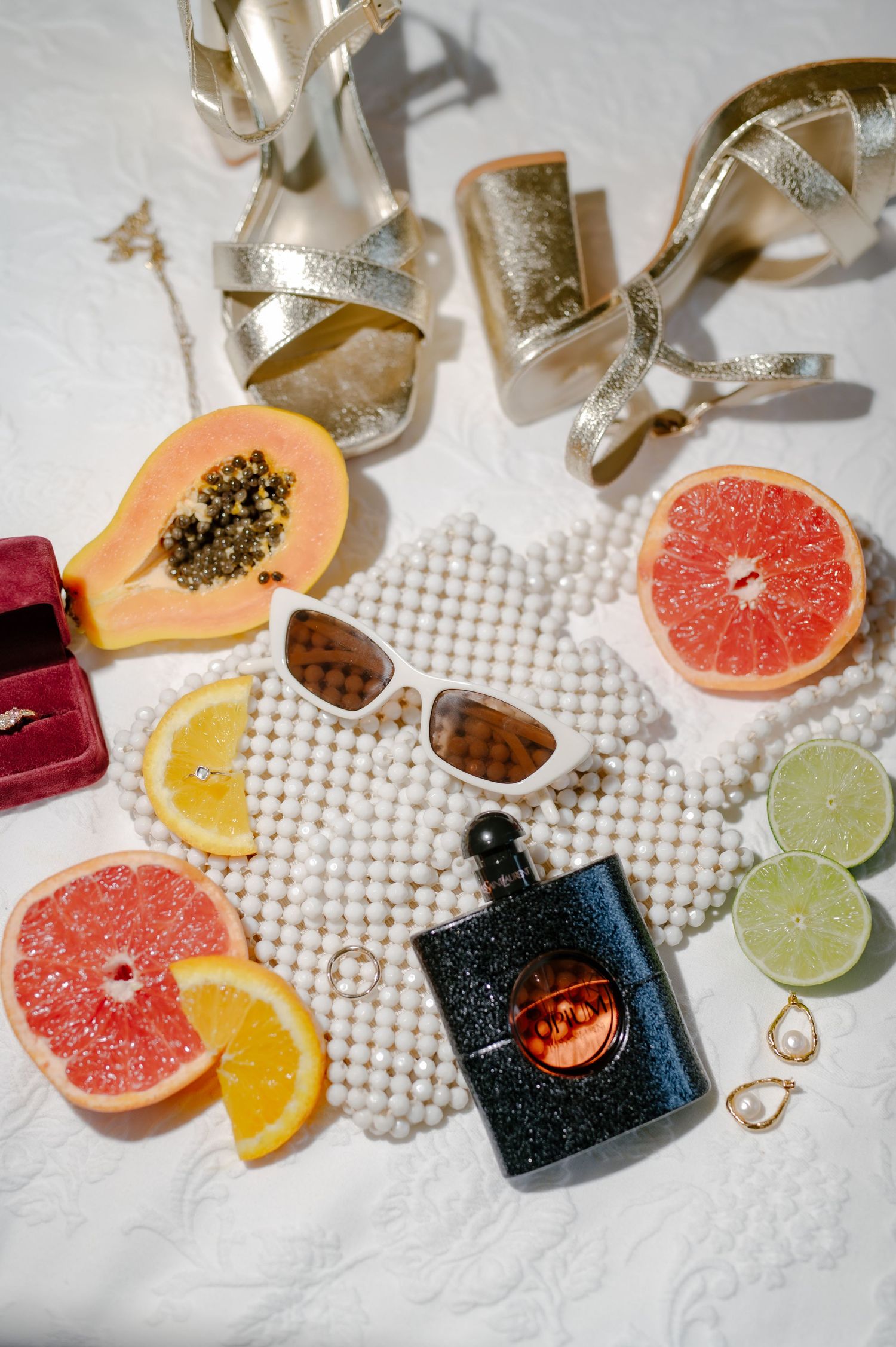 Tropical summer flatlay with citrus fruits, sunglasses, gold sandals, and perfume bottle on white textured surface.