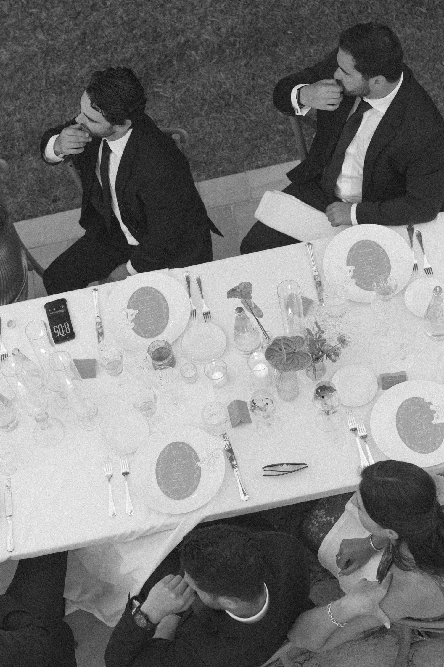 Aerial black and white photo of guests in formal suits seated at an elegantly set dining table at an event.
