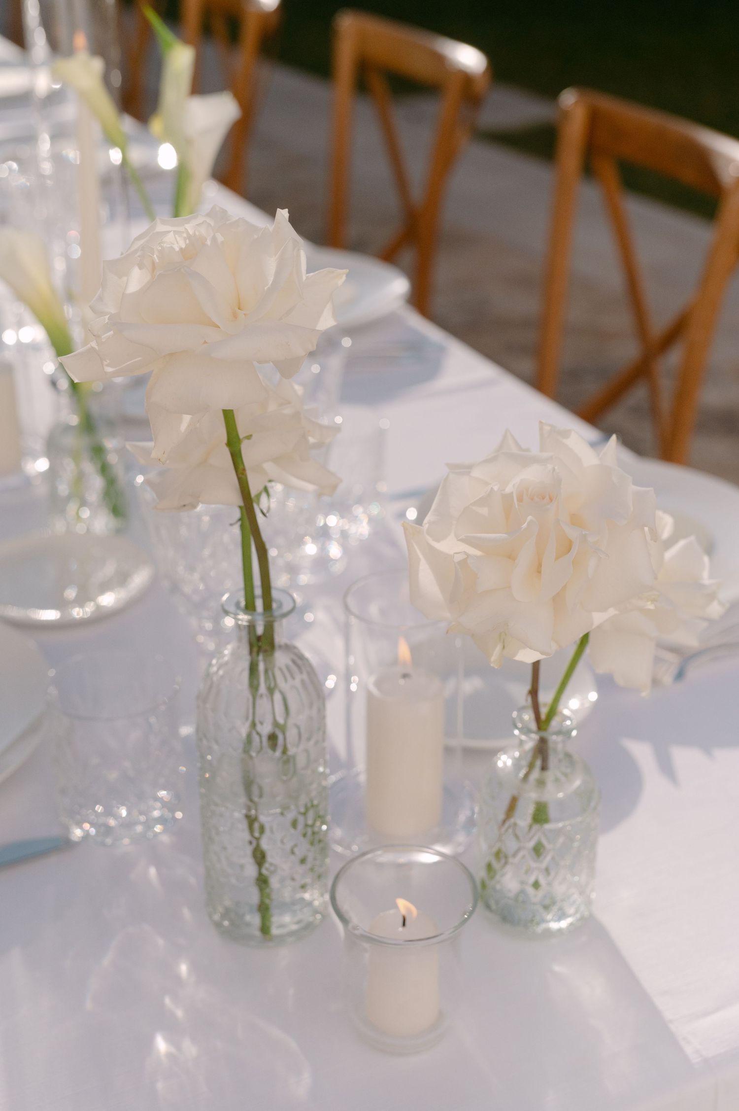 White roses in glass bud vases with pillar candles on a white linen tablescape for an elegant wedding reception.
