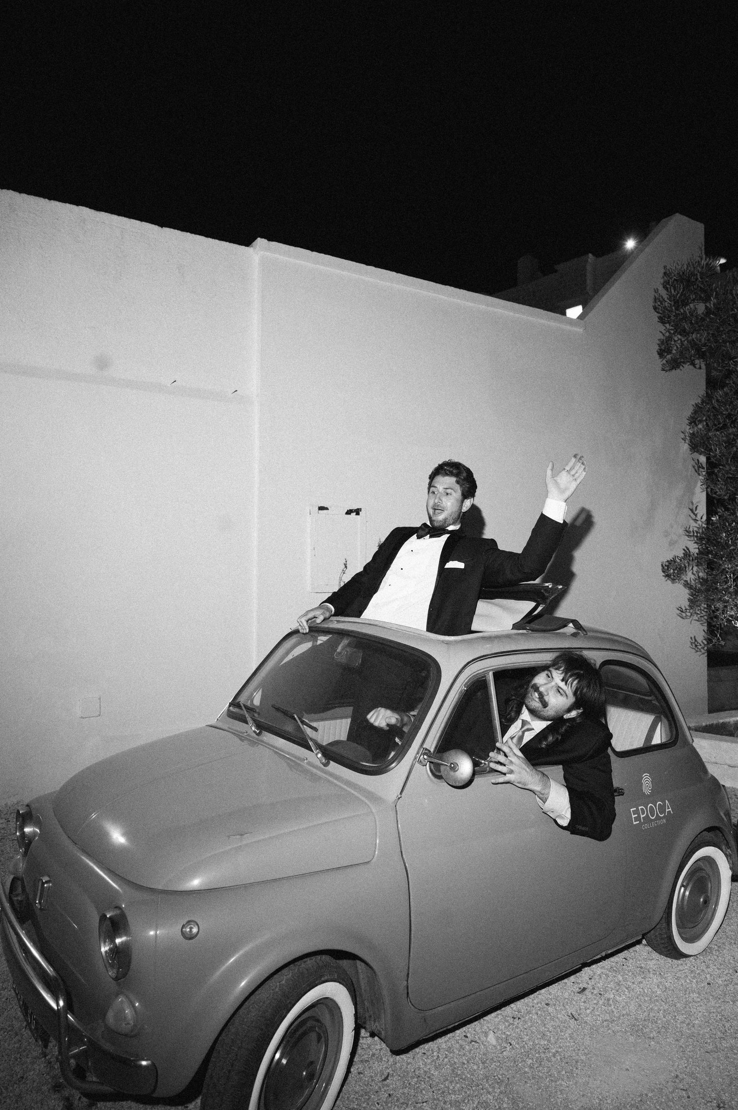 Two men in tuxedos celebrate atop a vintage Fiat 500 car in a black and white nighttime photo.