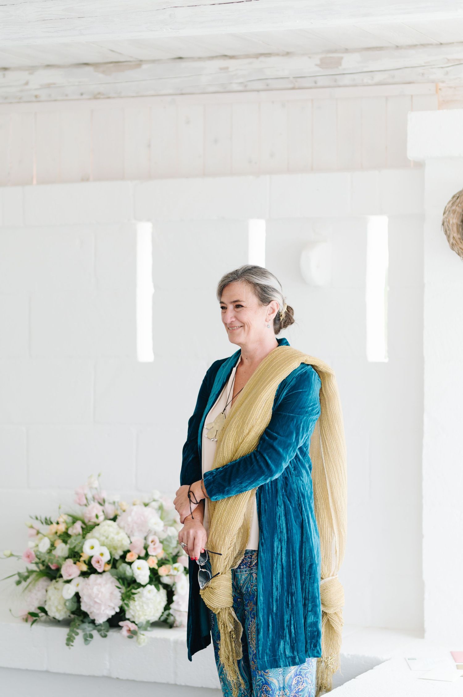 A person wearing a teal cardigan and yellow scarf stands in a bright white room with floral arrangements.