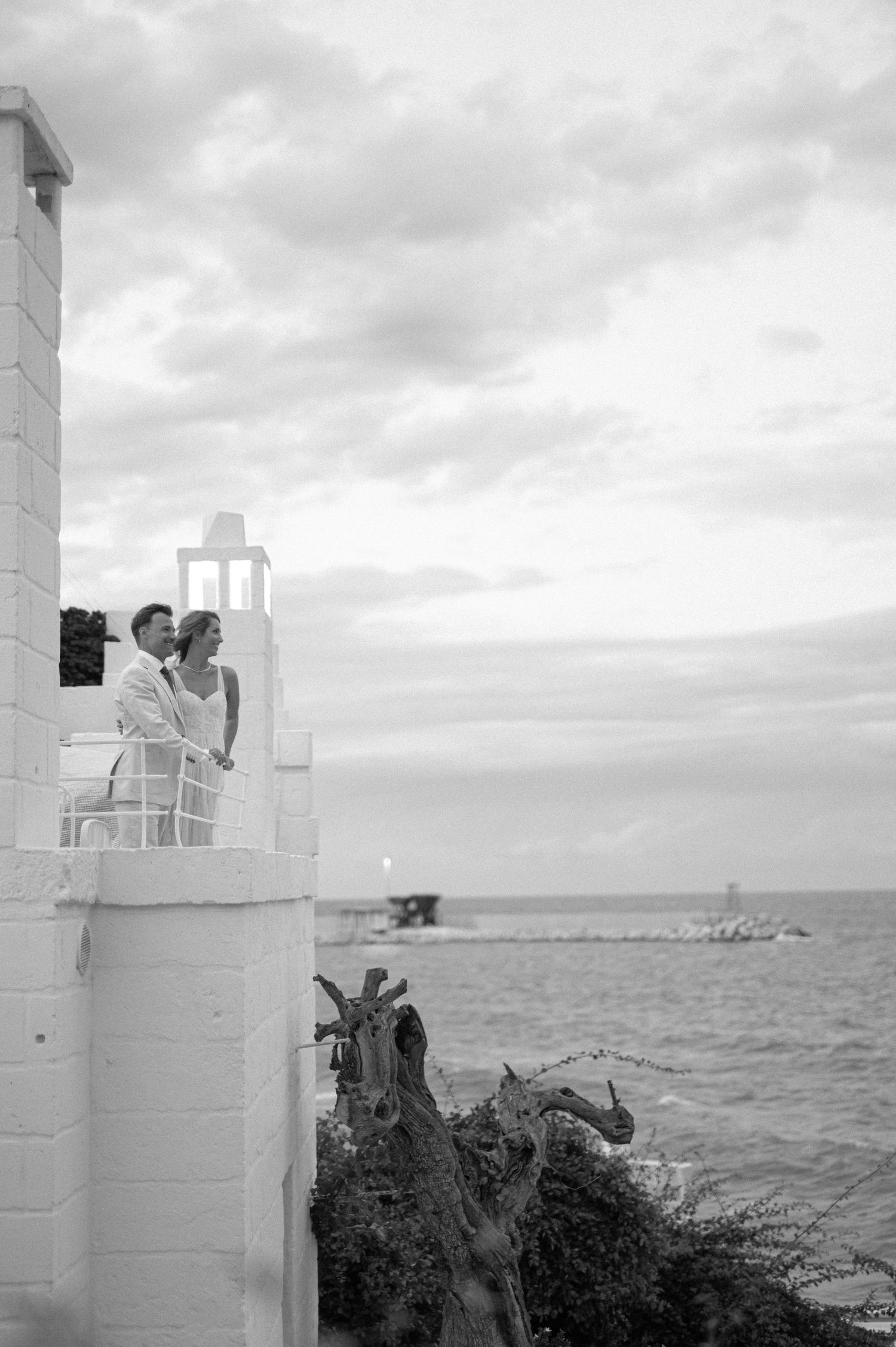 Black and white photo of two people embracing on a white lighthouse balcony overlooking the sea.