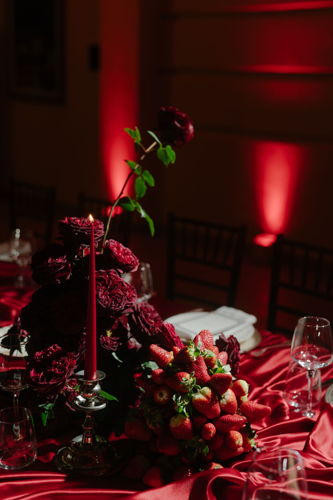 Fresh strawberries and raspberries arranged on a dark table with moody red lighting and glassware create an elegant display.