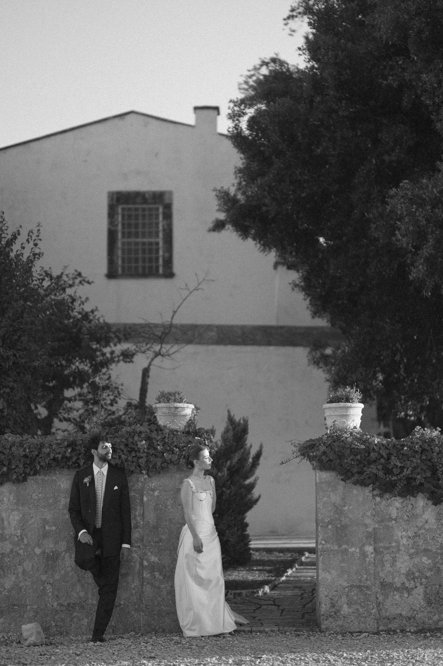 Black and white panoramic photo showing a couple walking along a stone wall in a garden setting.