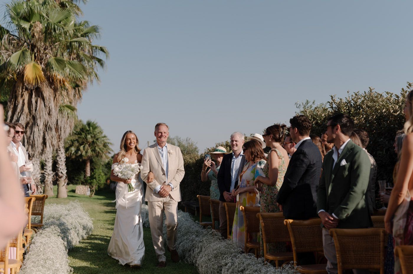 Guests seated outdoors at a wedding ceremony with palm trees and natural lighting.