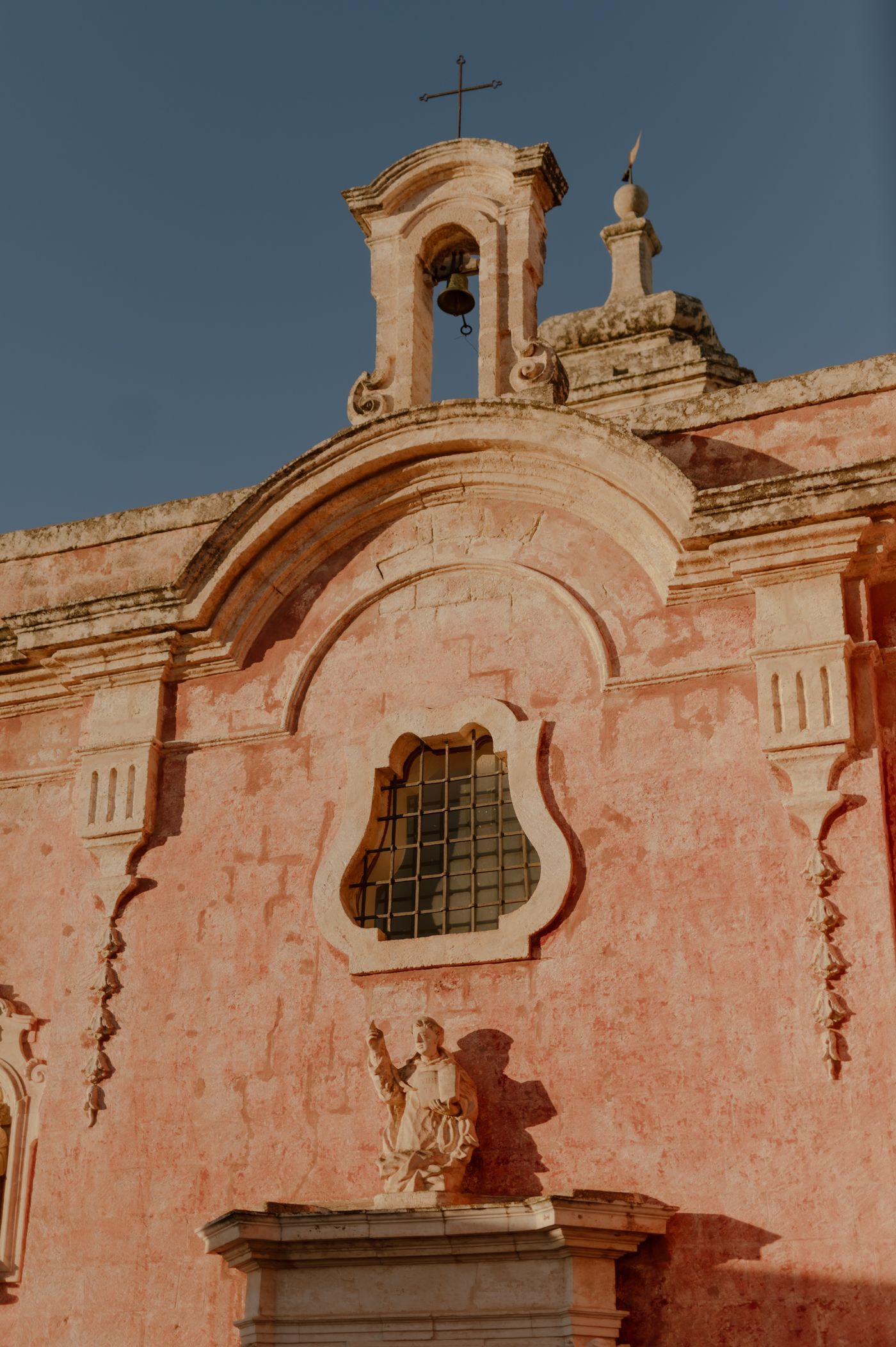 Pink stone church facade with ornate baroque architectural details and cross atop bell tower against blue sky.